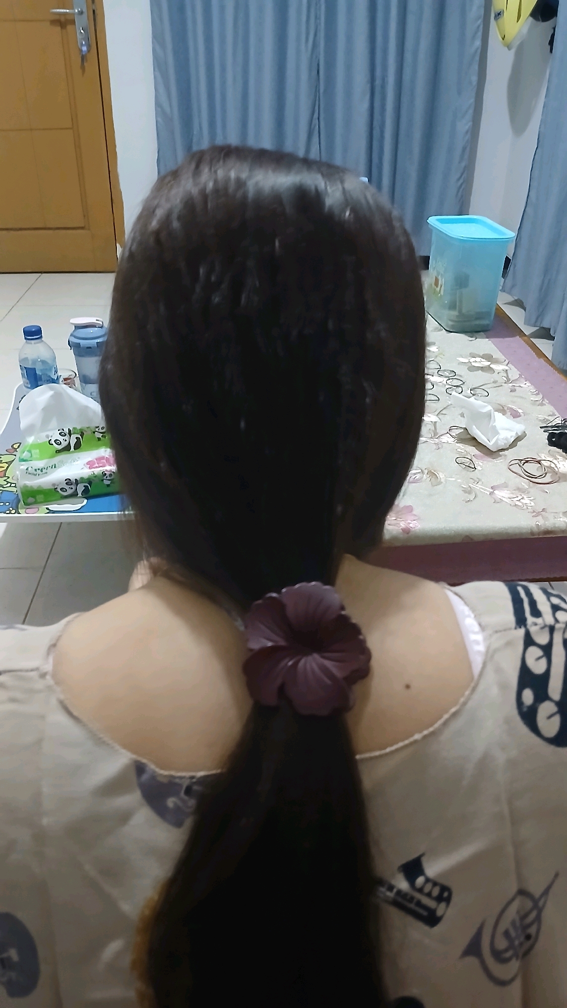 The image shows a close-up, rear view of a person with long, dark hair tied back with a dark purple scrunchie. The person is wearing a light-colored garment with a pattern that appears to include musical instruments. In the background, on a table, are several items including a box of tissues with panda designs, a water bottle, and a blue plastic container. A patterned tablecloth covers the table. To the left, a wooden door with a silver handle is visible. Blue curtains hang in the background. The environment suggests an indoor setting, possibly a home. The specific location in Sampang, Indonesia, is not identifiable from the visual information provided.