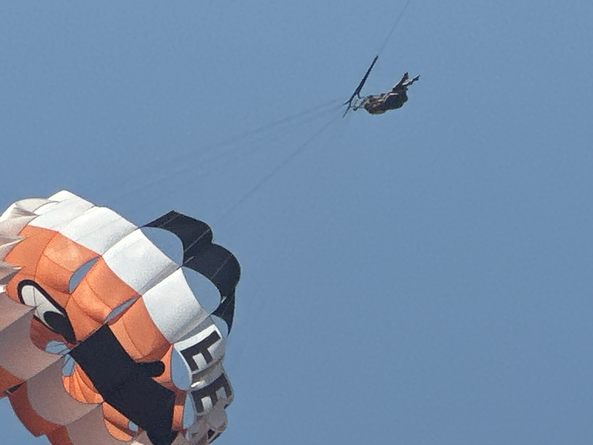 The image captures a parasailer suspended against a clear blue sky. The parasail itself is a large, colorful canopy with white, orange, and black sections, designed with a cartoonish face. Below, a person is attached to the parasail by ropes, appearing to be enjoying the flight. The scene suggests an activity taking place in an open, outdoor area, possibly a coastal region given the context of parasailing. While the precise location in Boreti, Montenegro is not explicitly visible, the activity of parasailing is commonly associated with tourist destinations and beaches.