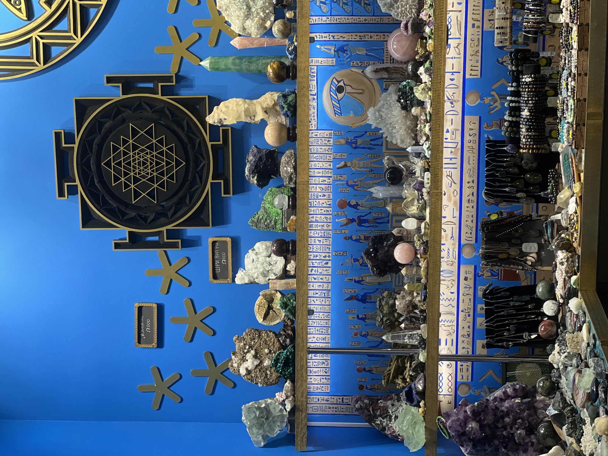 Here is a description of the image:
The image depicts a shop display, likely located in Moscow, Russia, given the Cyrillic text on the price tags. The wall is painted a bright blue and adorned with golden geometric symbols. Two framed signs display prices, one labeled "ИЗОБИЛИЕ" and the other "ШРИ ЯНТРА," both priced at 17,000. The display features shelves filled with a variety of crystals, minerals, and jewelry. The backdrop of the shelves is decorated with Egyptian-style hieroglyphics and imagery, painted in blue and white. On the wall, there are two distinct symbols in black and gold: one is circular with concentric geometric patterns, and the other is a square shape containing a complex triangular design inside a circle. Additionally, there are several golden, star-shaped symbols. The shelves hold a wide assortment of crystals, polished stones, and bracelets.