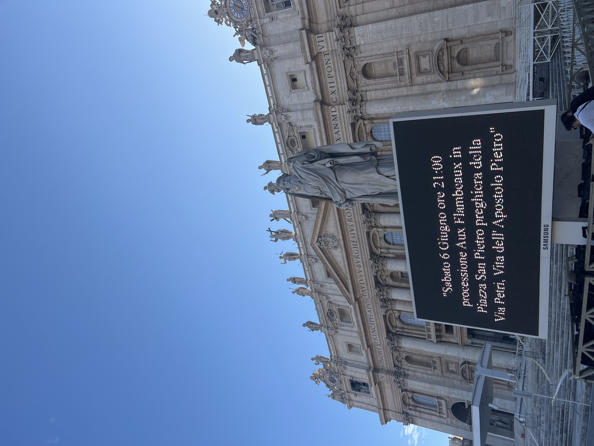 A low-angle, wide shot captures the grandeur of St. Peter's Square, Città del Vaticano, Italy, under a clear, bright blue sky. The upper two-thirds of the image are dominated by the cloudless expanse.
The prominent structure in the midground is the ornate, classical facade of St. Peter's Basilica, constructed from light-colored stone. Its design features multiple levels with large windows, Corinthian columns, and pilasters. Along the top balustrade, a series of statues of saints is visible. A large clock face is embedded into the facade on the left side. Latin inscriptions are clearly visible across the entablature, including "IN HONOREM PRINCIPIS APOST PAULUS V BURGHESIUS ROMANUS PONT MAX AN MDCXII PONT VII."
In the lower-midground, positioned in front of the basilica facade, stands a large, weathered stone statue of a bearded man draped in robes, holding what appears to be a staff or key.
A prominent feature in the lower central part of the image is a large, rectangular LED display screen, branded "SAMSUNG" at its base. The screen has a black background and displays white Italian text, which reads: "Sabato 6 Giugno ore 21:00 processione Aux Flambeaux in Piazza San Pietro preghiera della Via Petri, Vita dell' Apostolo Pietro".
The foreground shows parts of temporary structures, including white metal scaffolding, barriers, and tiered steps, suggesting preparations for an event. Two individuals are partially visible in the lower right corner, with one in a white shirt and the other in a dark shirt, both appearing to look down. The overall scene is brightly lit by daylight.