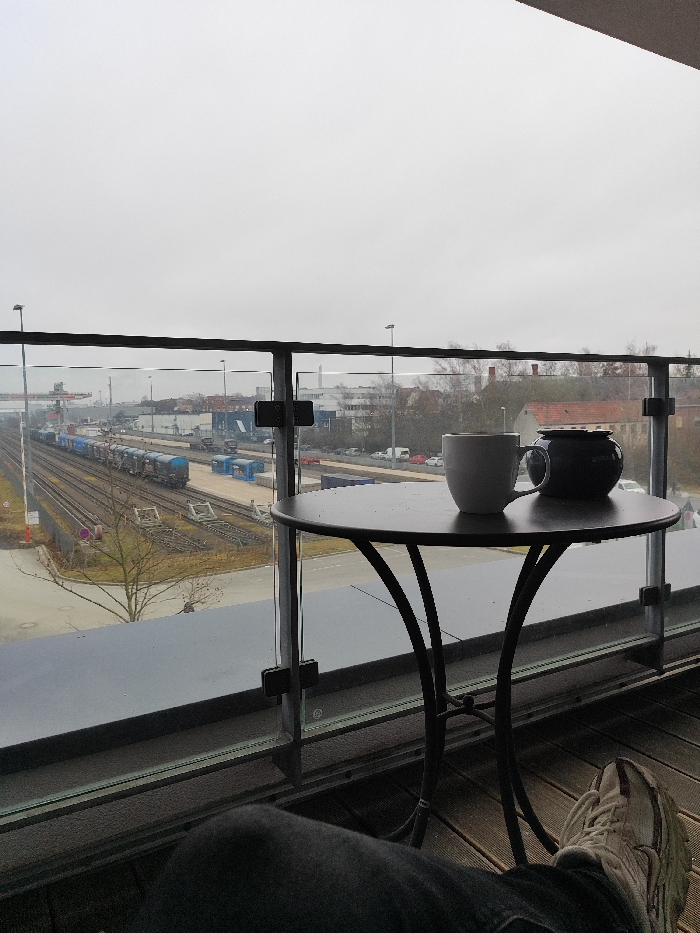 The image captures an elevated, static view from a balcony near Göttingen Hauptbahnhof, Göttingen, Germany, overlooking a railway yard and adjacent industrial area under an overcast sky. In the foreground, the lower legs and feet of an individual, wearing dark trousers and light-colored sneakers, are visible, indicating a seated position. A dark, round bistro table with curved metal legs is centered on the balcony, holding a white ceramic mug to the left and a dark blue lidded pot to the right. A glass and metal balcony railing extends across the mid-frame. Beyond the railing, multiple railway tracks traverse the scene horizontally from left to right. A stationary freight train with blue and grey wagons occupies one of the tracks. Parallel to the railway, a paved area features parked vehicles, including cars and trucks, and blue container-like structures. In the distant background, industrial buildings, a large crane, and sparse, leafless trees are discernible beneath a uniformly grey, cloudy sky.