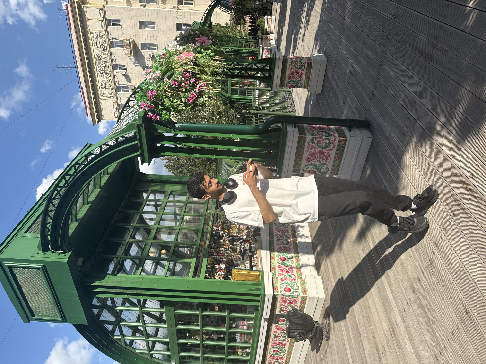 The image captures a sunny day at a pedestrian area featuring an elaborate green pavilion with arched glass windows and decorative floral accents. A young man, wearing a white t-shirt, black pants, and headphones around his neck, is looking at his phone while standing on a wooden boardwalk. In the background, a light-colored building with ornate architectural details stands under a clear blue sky with scattered clouds. The pavilion itself appears to be a shop or kiosk, with numerous small items displayed behind its glass facade. The scene is likely located in a public space or park within Moscow, Russia, possibly near a historical or cultural landmark given the architecture of the building and the style of the pavilion.
