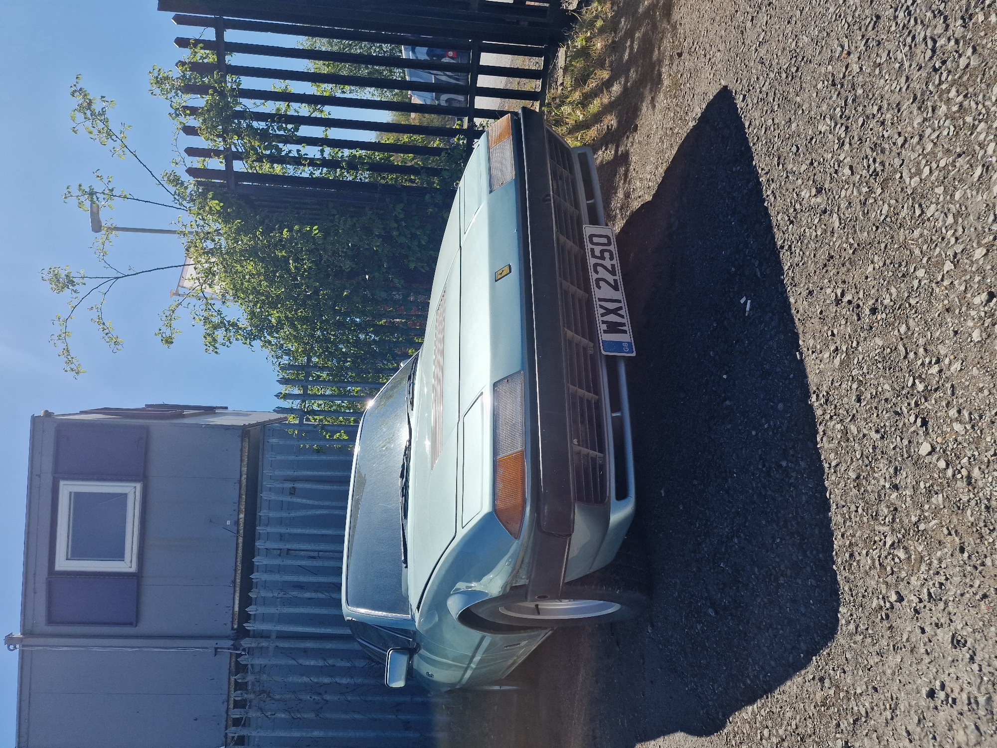 A light blue or teal classic car, possibly a Ferrari based on its angular design and small badge on the bonnet (though the badge itself is indistinct), is prominently featured in this outdoor image. The car is parked, showcasing its front and front-right side, with its polished surface reflecting the bright blue sky above. A UK license plate, clearly visible on the front, reads "WXI 2250" with the "GB" identifier.
The setting appears to be an industrial or commercial area in South Tyneside, United Kingdom, under bright, clear conditions. To the left of the car, a two-story structure resembling stacked portable cabins or shipping containers is visible, predominantly gray-blue, with a white-framed window on the upper unit. Behind the car and extending across the background is a dark metal fence, likely security fencing, entwined with green foliage and branches. The ground is a mix of gravel and asphalt, with a distinct, long shadow cast by the car, indicating strong sunlight, likely in the mid-morning or late afternoon. No people or specific activities are visible; the scene captures a static moment on a sunny day.