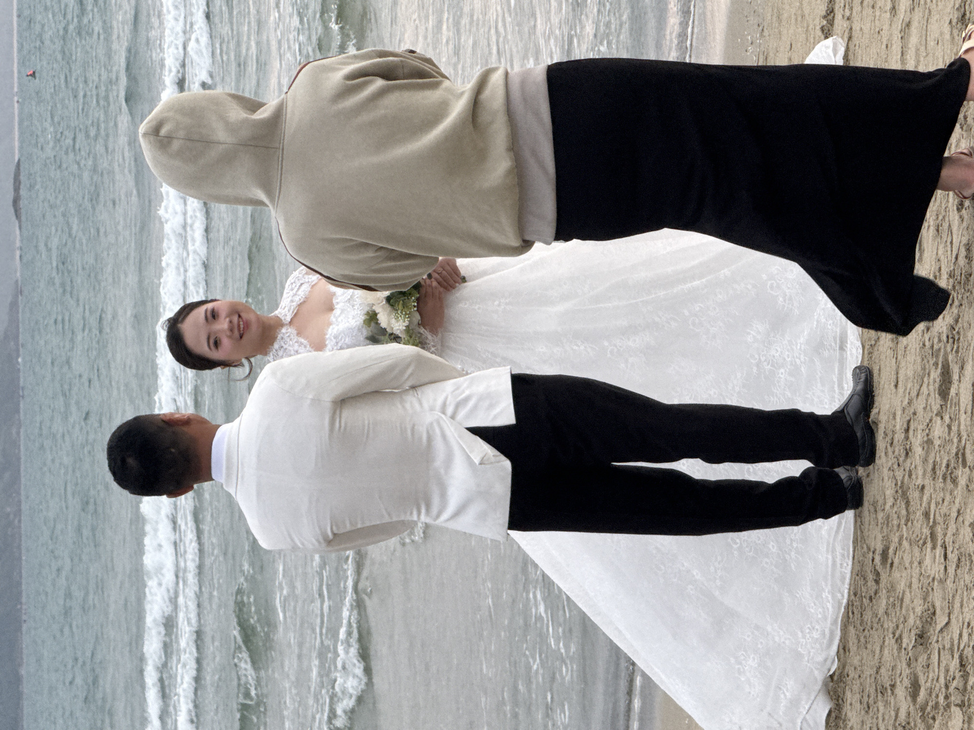 This image captures a serene wedding moment on a beach in Đà Nẵng, Vietnam. Three individuals are present. In the center, a smiling woman, presumably the bride, wears a white, lace-detailed strapless wedding gown and holds a bouquet of white and green flowers. She looks directly towards the viewer. Beside her, a man in a white suit jacket and black trousers, likely the groom, stands with his back largely to the camera, his black dress shoes visible on the sand.
Behind the couple, another person stands facing away from the camera, wearing a light beige hooded sweatshirt and a long black skirt, with light-colored sandals on their feet. This individual may be a photographer or assistant. The setting is a sandy beach with gentle, breaking waves in the background, extending into a calm, grey-blue ocean. The soft, natural light suggests an overcast daytime, contributing to a tranquil atmosphere. A small, distant object is visible in the water on the far left. No text is visible in the image.