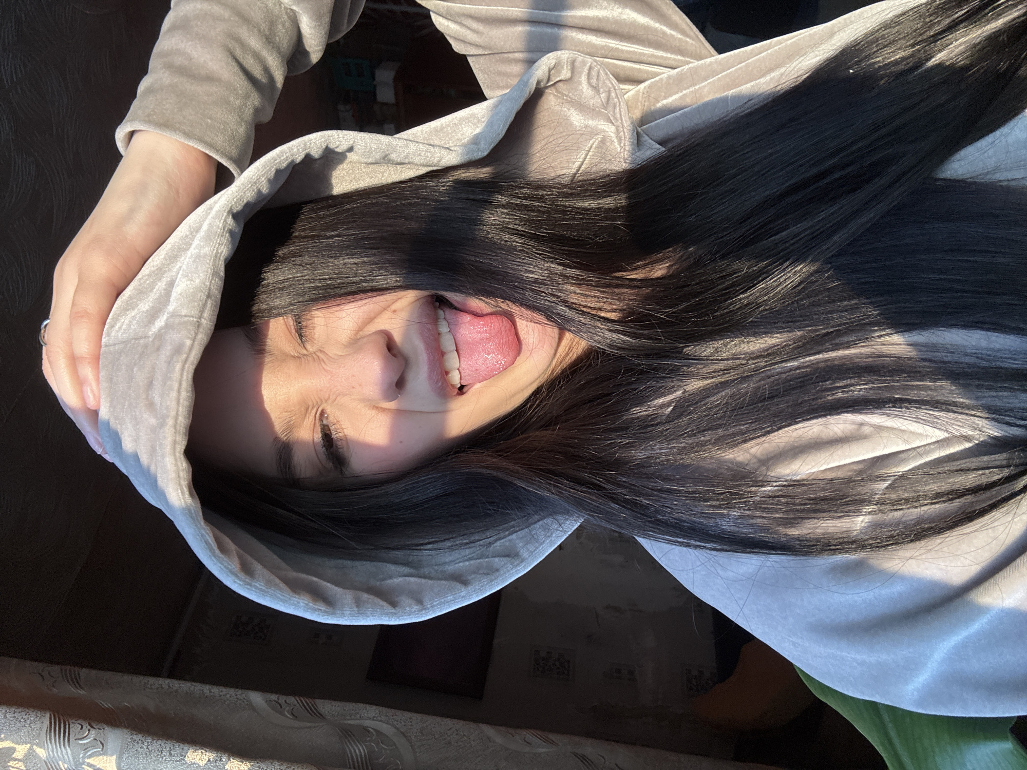 A woman wearing a grey hooded sweatshirt is sticking out her tongue. Her long black hair is cascading around her face and the hood. The setting appears to be indoors, with hints of domestic furniture and décor visible in the background, which is not clear enough to confirm the location as Фоки, Russia.
