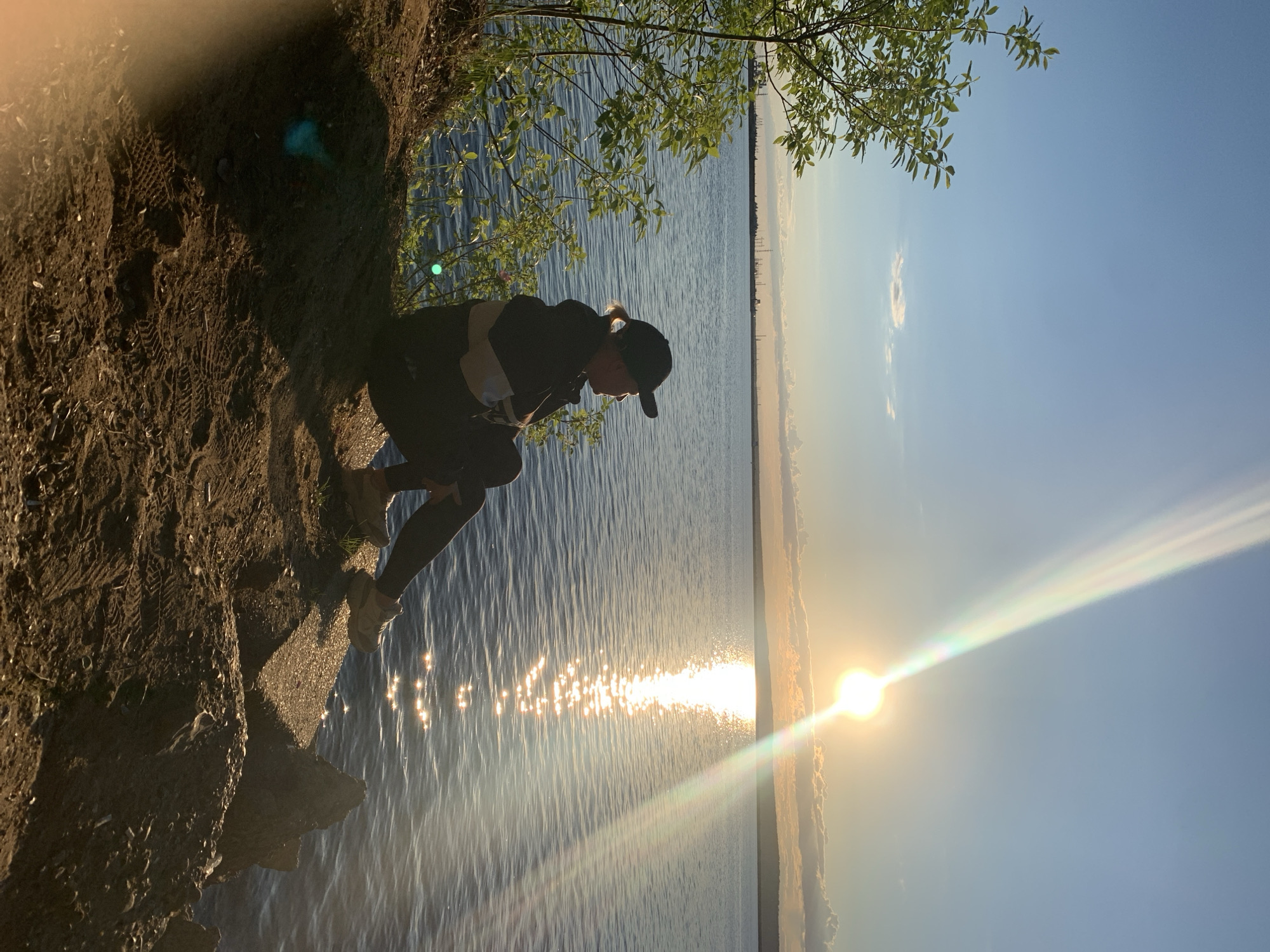 The image, set in Chaikovsky, Russia, captures a person crouching on a dirt bank overlooking a vast body of water, likely a lake or wide river, during sunrise or sunset. The sun is low on the horizon, casting a brilliant golden reflection across the rippled water and creating a prominent lens flare in the sky.
The subject, seen in profile, wears dark activewear including a cap, a long-sleeved top, and pants. They are positioned at the edge of the bank, looking intently towards the sparkling water. Their light-colored hair is pulled back in a ponytail beneath the cap. The bank itself is composed of dark, uneven earth with visible footprints, suggesting it's a well-trodden path. Sparse green vegetation and a tree branch with vibrant green leaves extend from the upper left, partially framing the scene.
The sky above is clear blue, with a faint line of distant land or clouds visible on the far horizon. The overall impression is one of peaceful observation or contemplation in a natural, open environment. No visible text is present in the image.