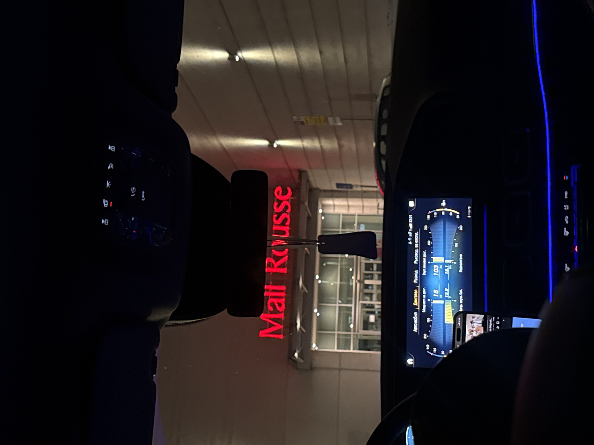 The image was taken from inside a vehicle at night, looking out towards the entrance of "Mall Rousse" in Ruse, Bulgaria. The mall's name is illuminated in red neon lights above the entrance. The interior of the car is dark, with blue ambient lighting visible on the dashboard and console. A car's digital display screen is in the foreground, showing various car metrics and a small phone screen displaying an image. A car air freshener hangs from the rearview mirror. The exterior of the mall appears modern with glass doors and windows. The overall scene depicts a typical urban nighttime environment.