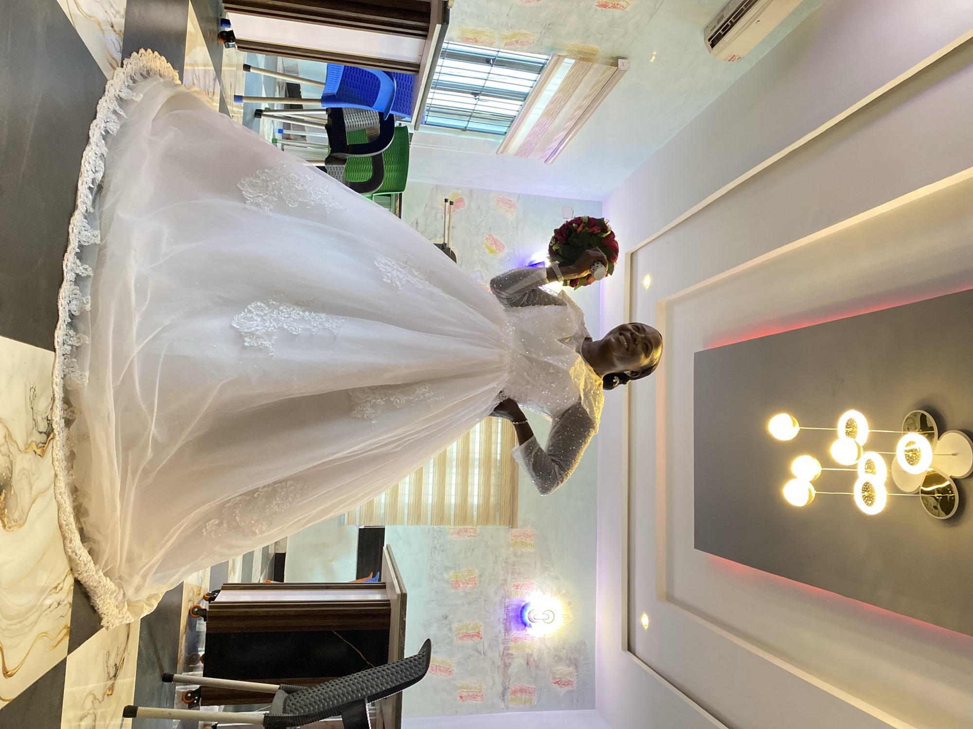 A woman in a white wedding gown is holding a red bouquet in a brightly lit interior. She is standing in a room with a black and white checkered floor, a wooden table, and blue and green chairs in the background. The walls have a decorative pattern, and a modern chandelier hangs from the ceiling. A window is visible in the upper part of the frame. The location is likely an indoor space, possibly a residence, in Kuru Dungus, Nigeria.