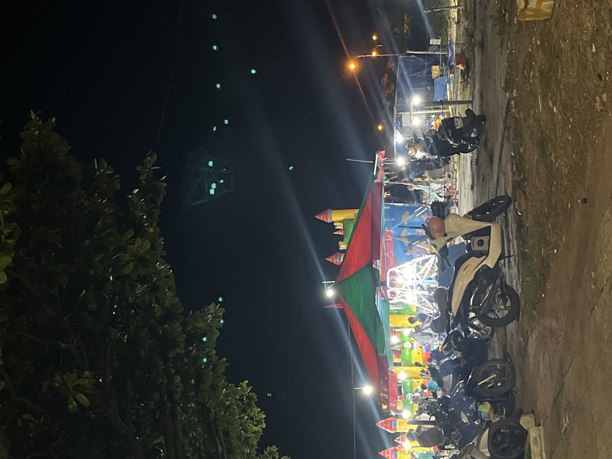 The image shows a nighttime scene in Bac Nha Trang Ward, Vietnam. A small fair or carnival is visible, with colorful tents, inflatable castles, and rides illuminated by bright lights. Several motorcycles are parked along the edge of the pavement next to the fair. People can be seen milling around within the fair area. A tree is visible on the left side of the frame, obscuring some of the background. In the dark sky above, there are some glowing objects that appear to be part of a Ferris wheel or other lit-up ride structure. The surrounding environment appears to be a street or open area with a rough, unpaved section along the right side.