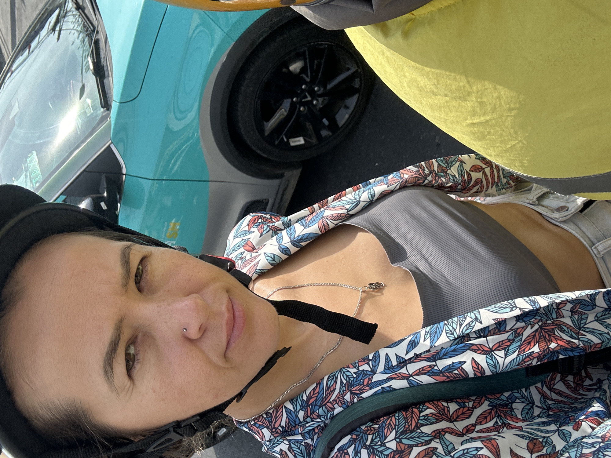 A woman wearing a helmet and a floral-print shirt over a grey crop top is taking a selfie. She has a nose piercing and is wearing a silver necklace with a small pendant. In the background, a turquoise car is visible, along with the wheel and part of the door. The scene appears to be outdoors on a paved surface, possibly a street.