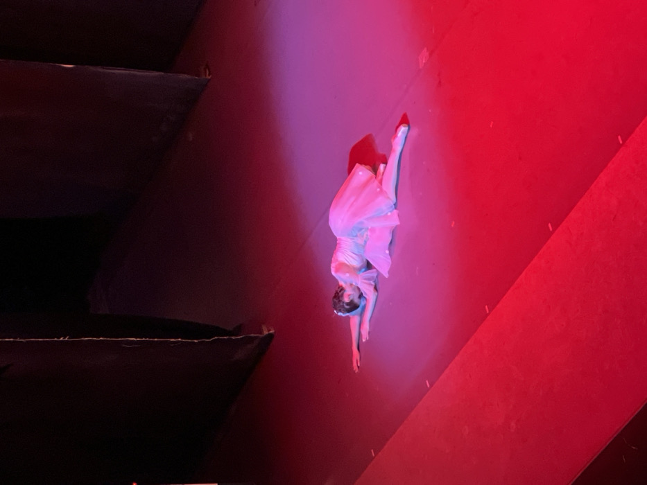 A ballerina, bathed in pink and red light, performs on a dark stage. She wears a pale dress and is in a split position. Her left leg extends upward and to the right, while her body and extended right arm stretch downwards. A dark, undefined structure looms on the left side of the frame, contrasting sharply with the bright stage lighting illuminating the performer.