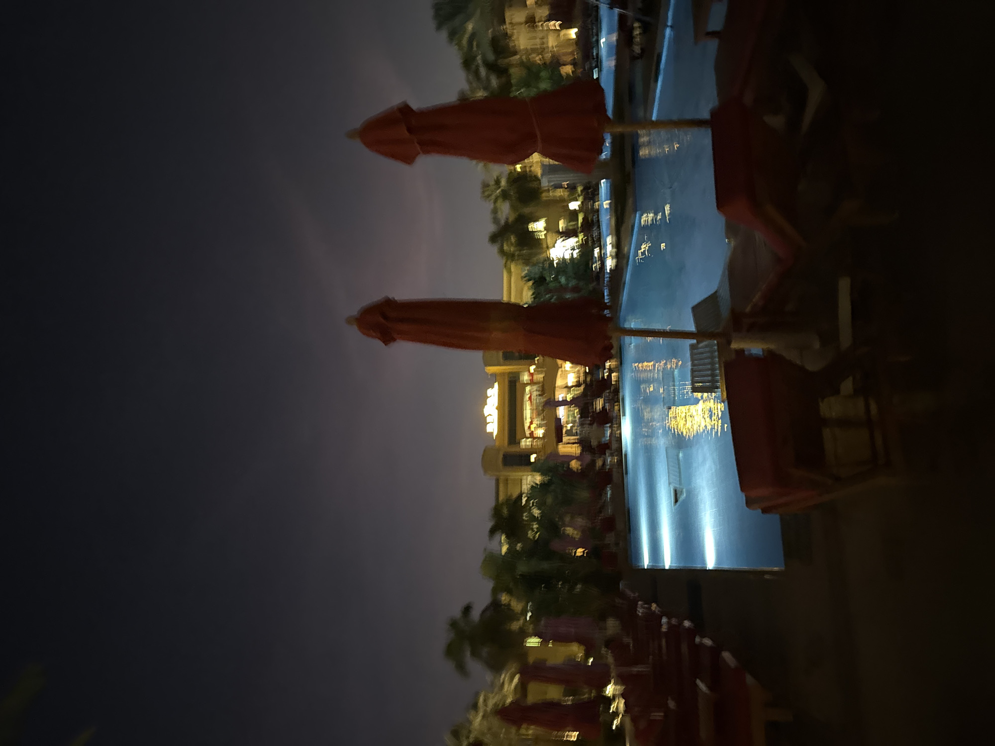 This is a night-time, wide-angle, blurred outdoor shot of a resort-style swimming pool area. In the foreground, several red lounge chairs are visible, some with red umbrellas open. The swimming pool, illuminated by underwater lights, occupies the central portion of the image, reflecting yellow lights from the buildings. In the background, a large, brightly lit building with an Arabic-style architectural design is visible. Palm trees are scattered throughout the scene. The sky is dark, with a faint gradient of lighter shades towards the horizon, suggesting dusk or early dawn. The location is identified as Unknown, Egypt.