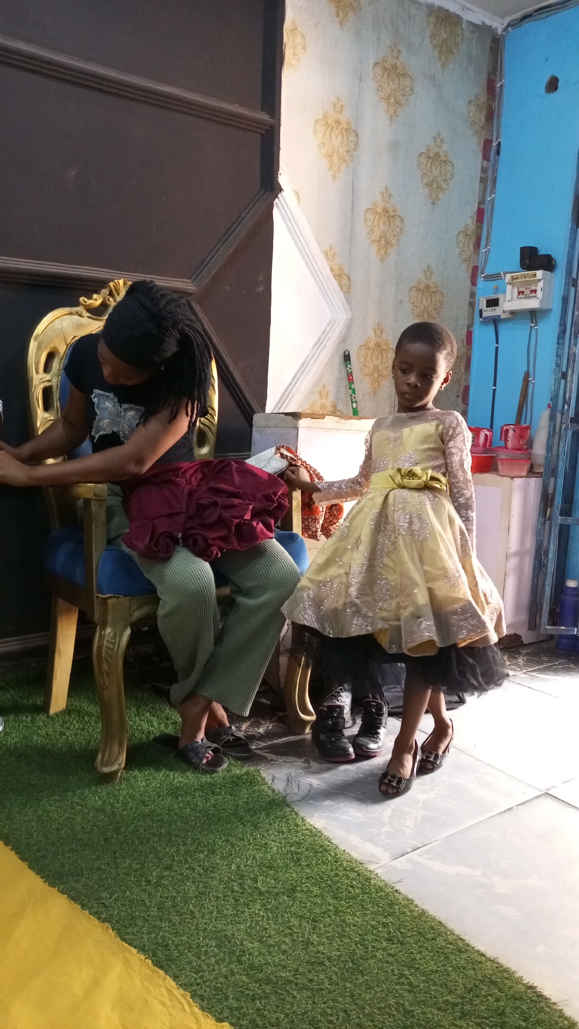 A young girl wearing a gold dress and oversized black high heels stands next to a seated woman. The woman sits on a blue and gold decorative chair, holding a red garment. The girl holds a scarf with a floral design. A green artificial grass rug covers the floor in the left corner. The floor near the child is tiled. Behind the subjects is a wall with floral wallpaper. In the right corner, a wall is painted light blue, and there's an electrical panel mounted on it, along with some containers on a small white table. Based on the prompt's instructions, this image was taken in Sabon Tasha, Nigeria.