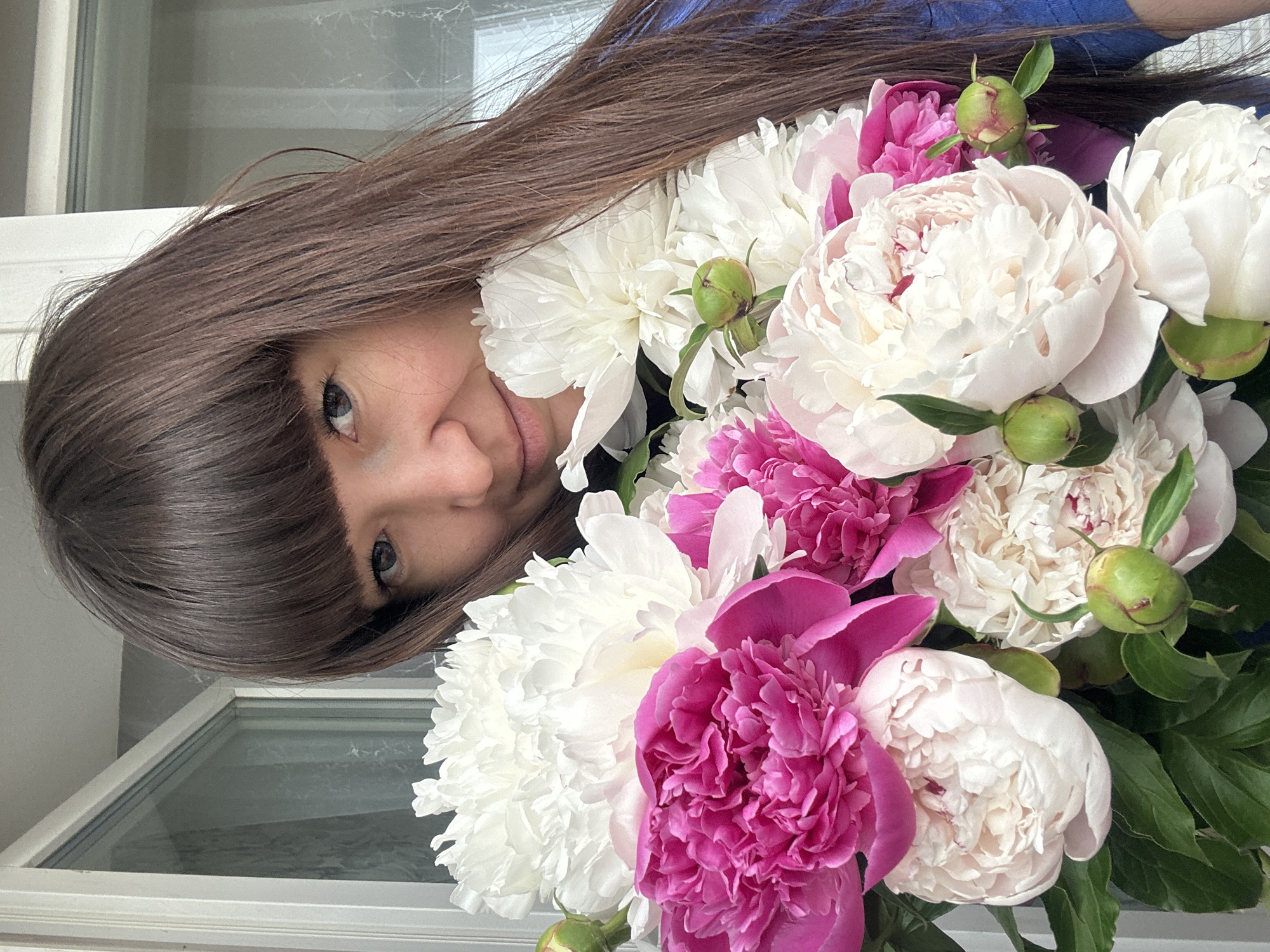 A woman with brown hair and bangs holds a large bouquet of white and pink peonies. She is looking at the camera with a slight smile. The scene appears to be indoors, near a window. There's no information suggesting this image was taken in the city of Chaikovsky, Russia.