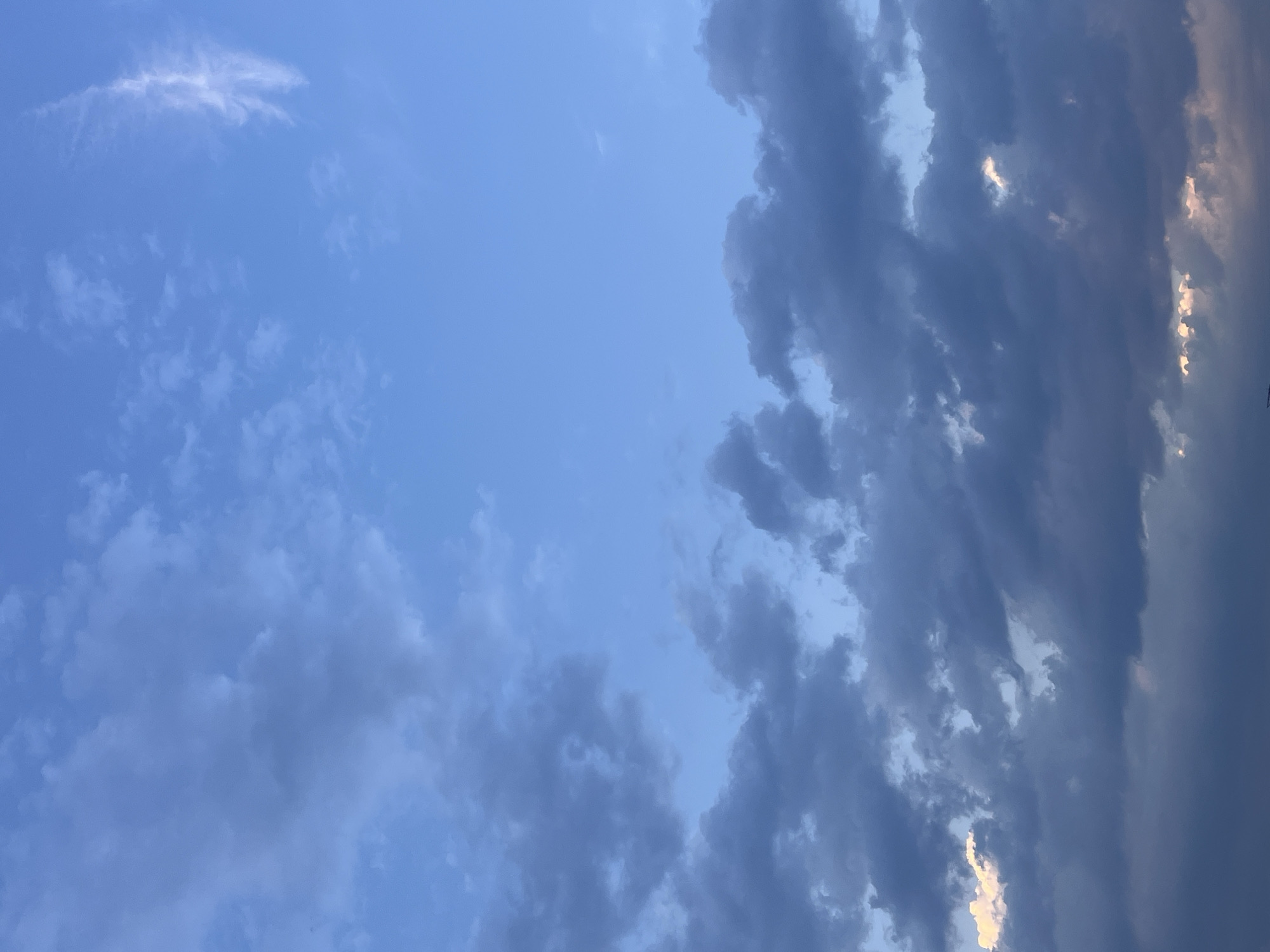 The image depicts a daytime sky over บ้านกาญจน์กนก 23, Thailand. The sky is primarily blue with scattered clouds. The clouds vary in density, ranging from thin wisps to denser, darker formations. Some areas of the clouds appear to have sunlight filtering through them, creating a brighter edge.