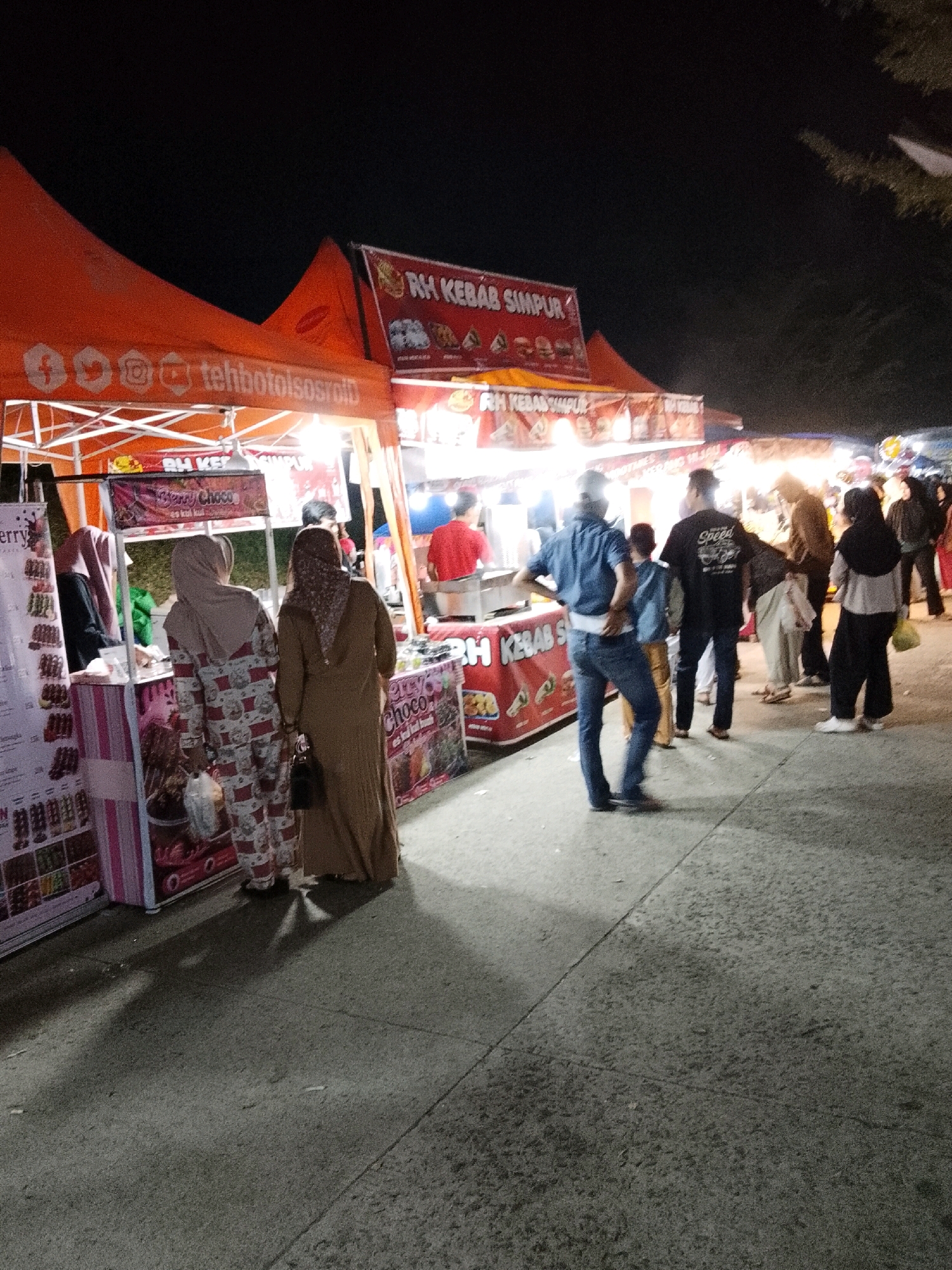 In the city of Padang Dalom, Indonesia, at night, a street food stall is depicted. The stall is adorned with an orange tent and features signs advertising "RH KEBAB SIMPUR". Several individuals are visible around the stall, some appearing to be customers and others possibly vendors. One person is wearing a blue shirt and jeans, another a black t-shirt with "Speed" written on it, and a few individuals are seen in hijabs. The ground is paved, and shadows are cast by the lighting, suggesting it is evening or nighttime. The scene portrays a typical bustling atmosphere of a local night market.