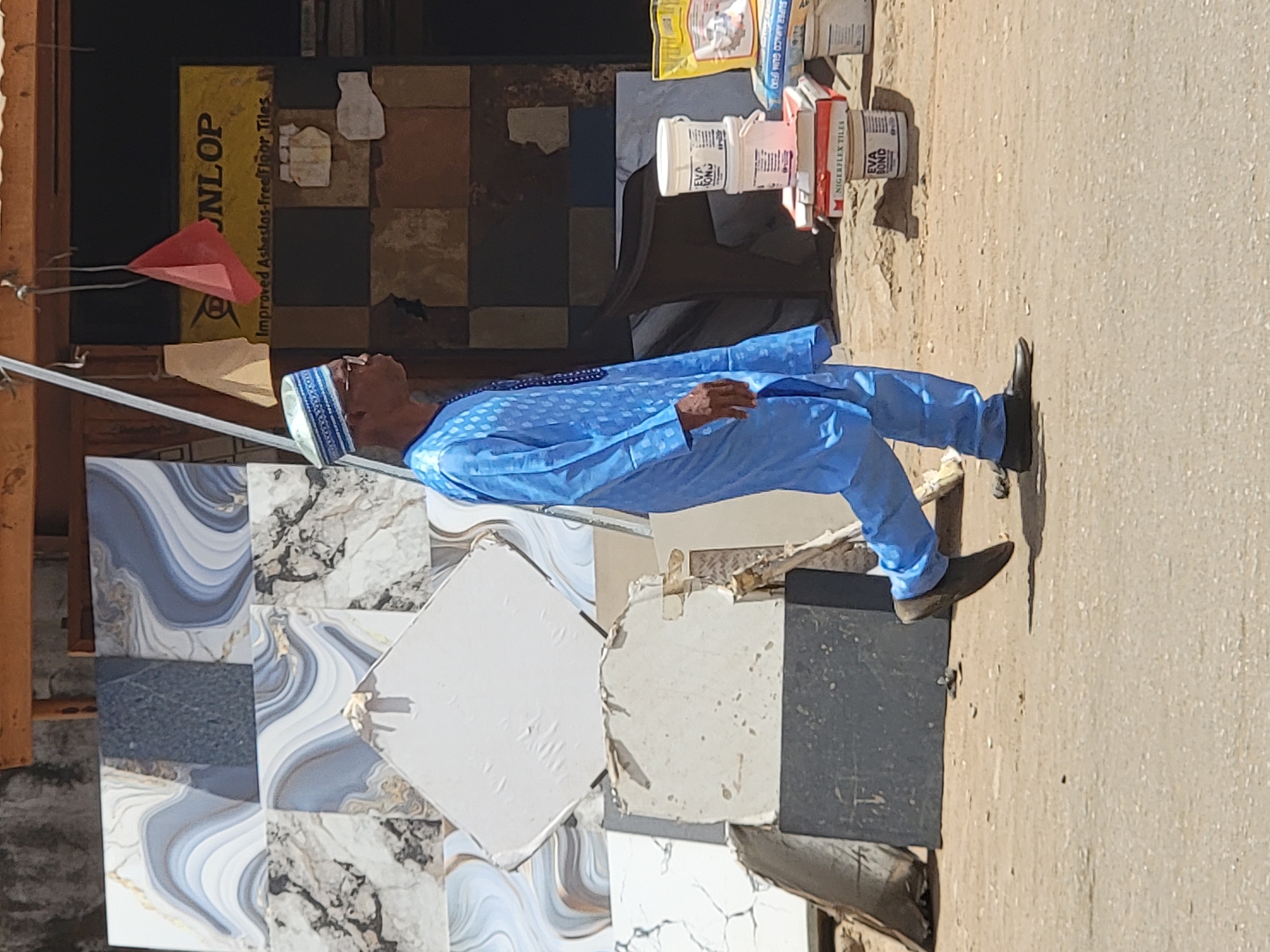 The image depicts a man in a light blue boubou and a kufi cap standing in front of a display of ceramic floor tiles. He has black shoes and is facing left, slightly angled towards the viewer. The tiles on display vary in color and pattern, including marble-like designs and solid colors like white and gray. Behind the tiles, there are construction materials, including cans and bags of adhesive and tile grout, as well as a banner that reads "JNLOP Improved Asbestos-free floor Tiles." The ground appears to be a dusty, paved area. The location is indicated to be in Maiburiji, Nigeria.