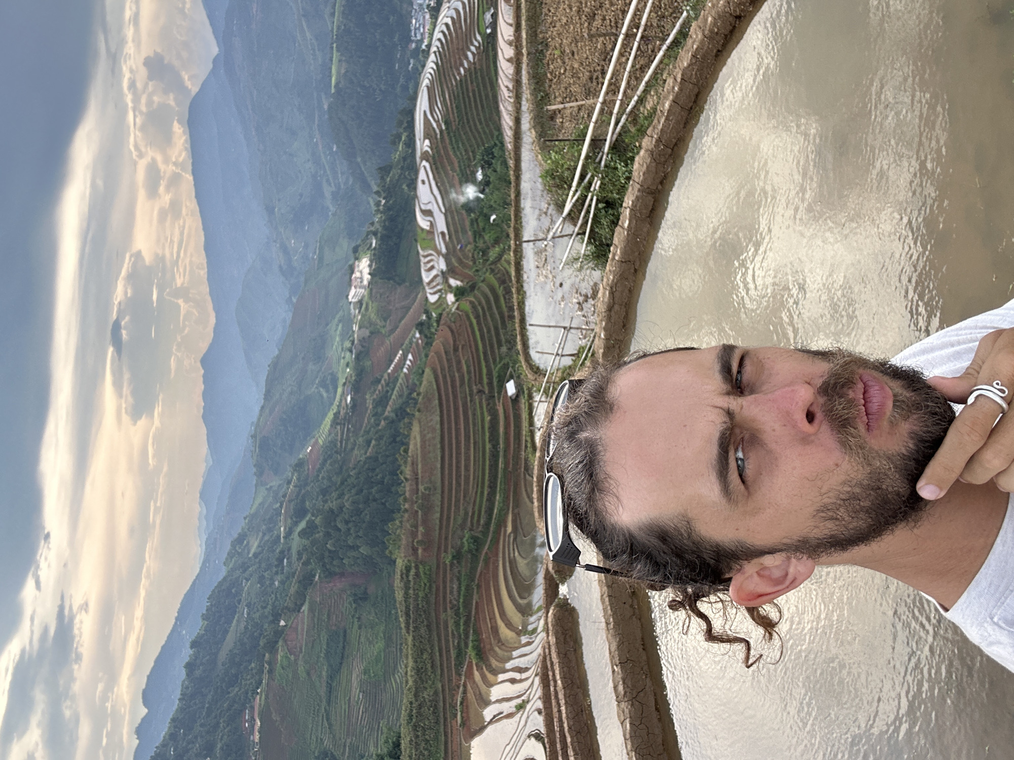 A portrait-style image captures a man with a beard, mustache, and dark curly hair, positioned on the right side of the frame. He wears a white t-shirt, with sunglasses perched on his head and a silver ring on his finger. His expression is serious, with one eye slightly squinted.
The background is a breathtaking mountainous landscape, characterized by extensively terraced rice paddies. Many terraces are filled with water, reflecting the cloudy sky and creating intricate patterns across the hillsides. A dirt path and some white poles are visible amidst the cultivation. Layers of mountains recede into the distance, partially obscured by atmospheric haze under a dynamic sky. The lighting, with hints of golden hues on the clouds, suggests either late afternoon or early evening. This scene depicts the man amidst the iconic cultivated terrain of places like Lao Chai, Vietnam. No text is visible in the image.