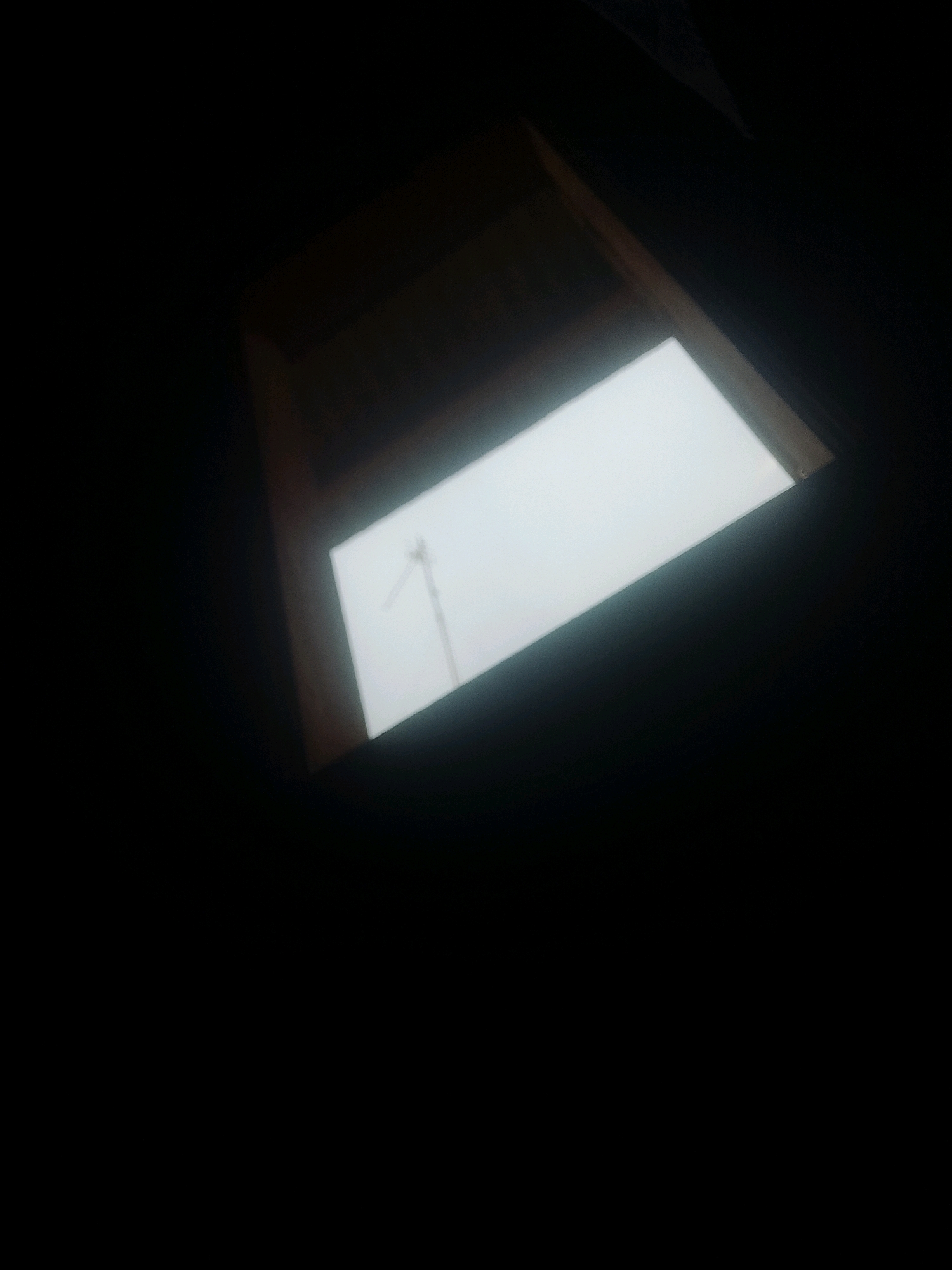The image shows a skylight with a view of a crane against a bright, overcast sky. The skylight is framed by dark wooden beams and appears to be in a dark, indoor space. The overall impression is one of looking up from within a building towards the outside world, a stark contrast between interior darkness and exterior light. The scene is devoid of people, and the only discernible object outside is the crane, suggesting it might be an urban or industrial setting. The location within Kecamatan Haurgeulis, Indonesia is not visually discernible from the image itself, and therefore cannot be included in the forensic description.