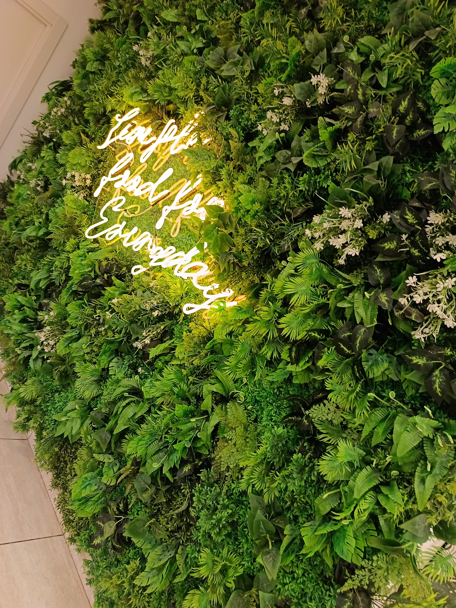 The image depicts a vertical garden wall, densely covered in a variety of green plants and small white flowers. A neon sign in a cursive font, emitting a yellow light, is mounted on the left side of the wall. The wall appears to be indoors, likely in a restaurant or commercial establishment. The photo's perspective is slightly angled. The wall's surface is textured with different leaf shapes and textures.
The location in Osa, Russia, is not identifiable from the image.