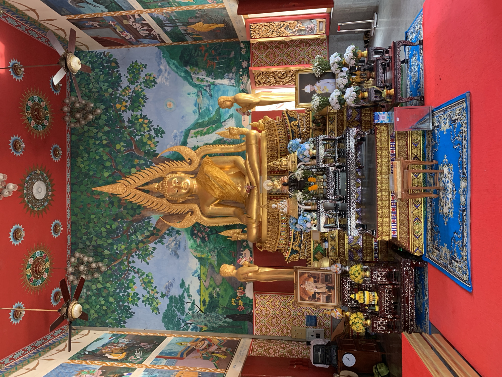 The image depicts the interior of a Buddhist temple on the island of Ko Samui, Thailand. The focal point is a large, ornate golden Buddha statue seated on a multi-tiered platform. Flanking the Buddha are two smaller golden statues. The wall behind the Buddha is adorned with a vibrant mural depicting a lush landscape with trees, mountains, and a body of water. On either side of the main Buddha statue are framed portraits, likely of royalty or revered figures, and decorative floral arrangements. The ceiling is painted red with intricate circular motifs and features two ceiling fans. Red carpeting covers the floor, with a blue rug placed in front of the altar area. Several wooden cabinets and tables with various offerings and religious items are visible around the altar. The overall atmosphere suggests a place of worship, with rich cultural and religious symbolism evident in the decor and objects.