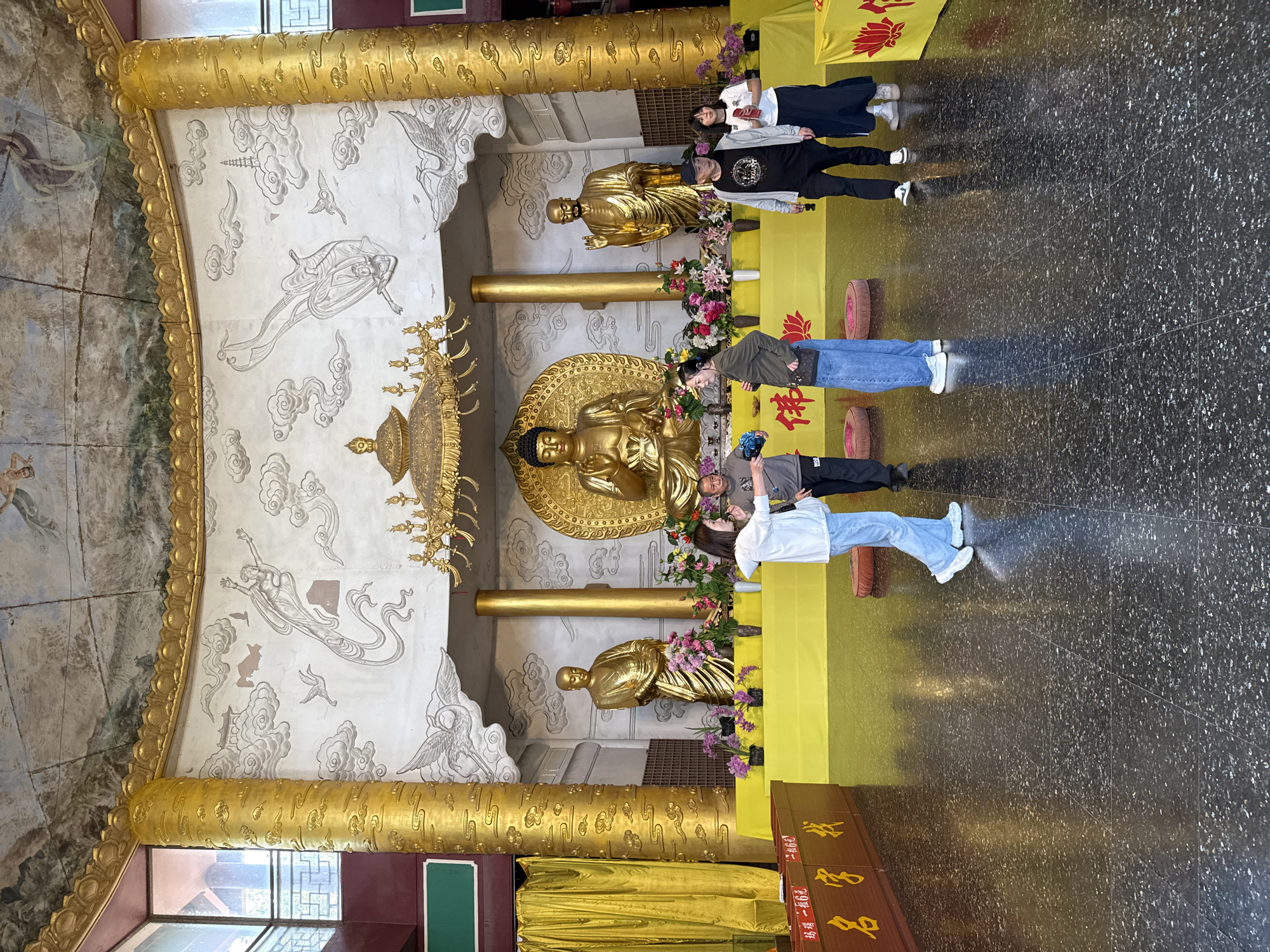 The image depicts a scene inside a temple, possibly located in Shizhong, China, where several people are standing near a display featuring three golden Buddha statues. The central, larger statue is seated, while the other two are standing. The statues are adorned with colorful flowers at their bases. Behind the statues, a yellow cloth backdrop hangs. The walls and ceiling are decorated with intricate golden designs and murals depicting figures and clouds. The floor is dark and glossy. A group of four people, two women and two men, are standing, seemingly posing for a photograph.