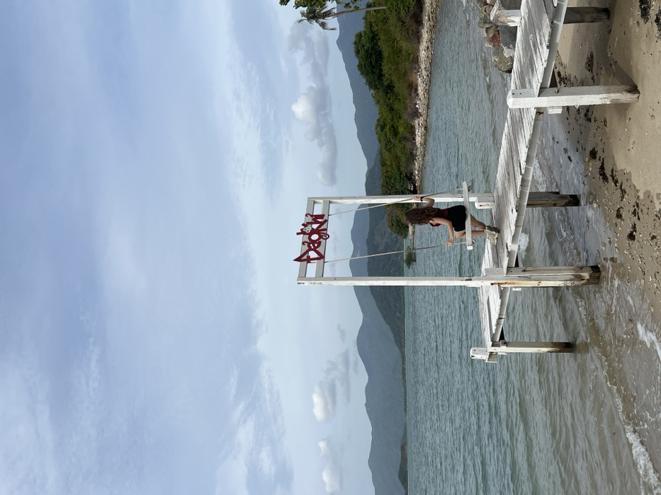 A coastal scene in Ninh Ích, Vietnam, depicts a weathered white wooden pier extending from a sandy and pebbly beach into a calm body of water under a partly cloudy sky. An adult female, with long, curly dark hair, is seated on a light-colored wooden swing suspended from a white wooden archway positioned on the pier. She wears a dark red top, a black skirt, and light-colored footwear, with her back to the viewer, gazing towards the distant horizon. Her hands grasp the swing ropes. The archway features a red sign at its apex displaying stylized text and two small figures. The background consists of a greenish-blue bay, a lush green shoreline to the right, and a range of dark green mountains beneath an overcast sky with scattered white clouds. The foreground beach contains sand, small pebbles, and scattered dark organic material.