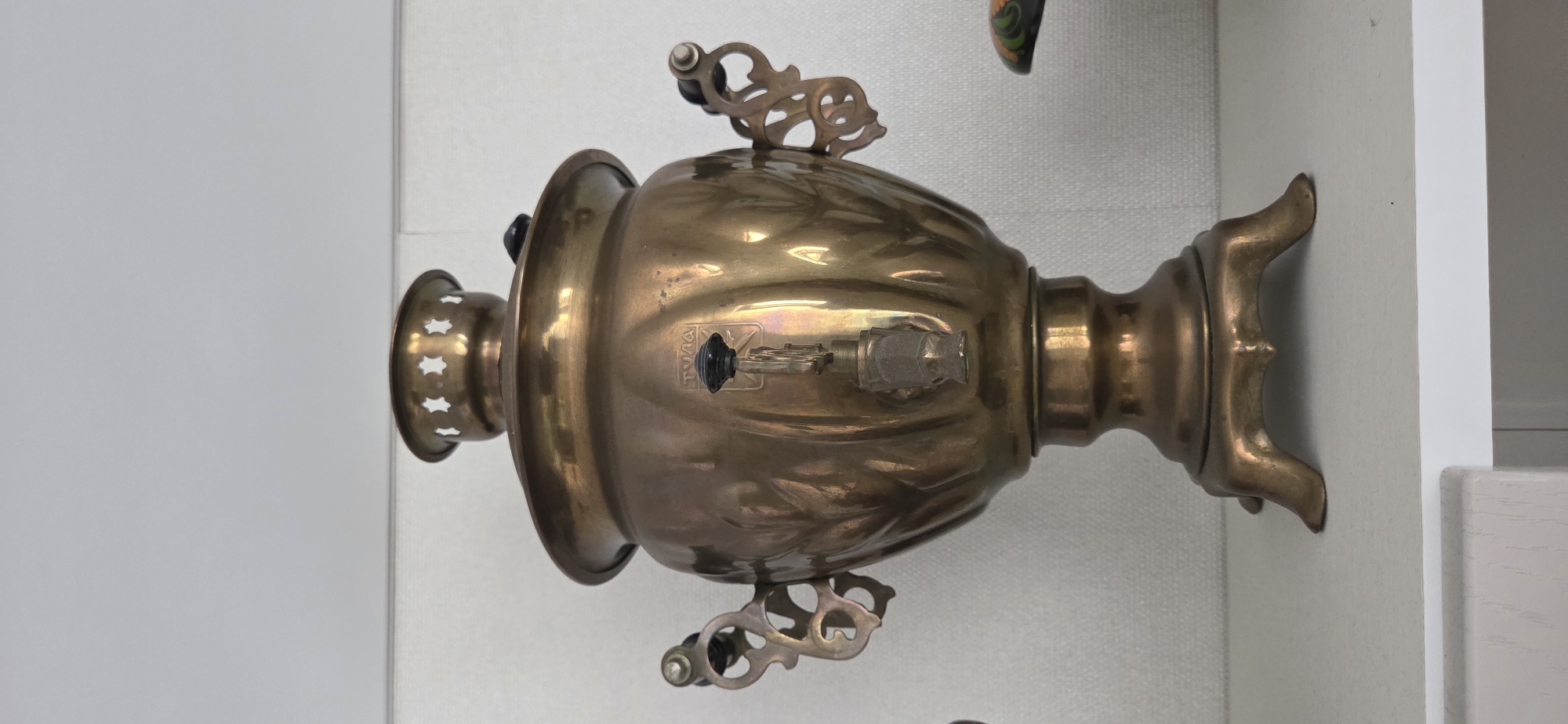The image displays a vintage brass samovar, rotated 90 degrees counter-clockwise, against a plain, light-colored background. No people or active events are depicted; the samovar appears to be stationary, possibly on display.
The samovar is crafted from polished brass, exhibiting a golden hue with some reflections and areas of slight tarnishing or patina. Its body features a classic, ornate shape, possibly with subtle engraved or embossed patterns. Two elegantly scrolled handles, each with a dark grip, extend from the sides of the main body. A spigot is prominently featured on the front, equipped with a black handle and a transparent, faceted knob. Above the spigot, an emblem or logo is visible, and the Cyrillic word "ТУЛА" (TULA) is clearly legible on the spigot mechanism, indicating its origin.
The samovar rests on a decorative base with four curved feet. The top section, which would typically hold a teapot or serve as a chimney, is visible on the left side of the rotated image, characterized by a perforated, decorative rim.
The background is simple, consisting primarily of a light grey wall or surface. A stark white vertical structure, possibly a doorframe or piece of furniture, is present on the right side. A small, colorfully painted object, resembling a decorative spoon or part of a souvenir, is partially visible and cropped at the top right corner. The lighting is consistent with an indoor setting.