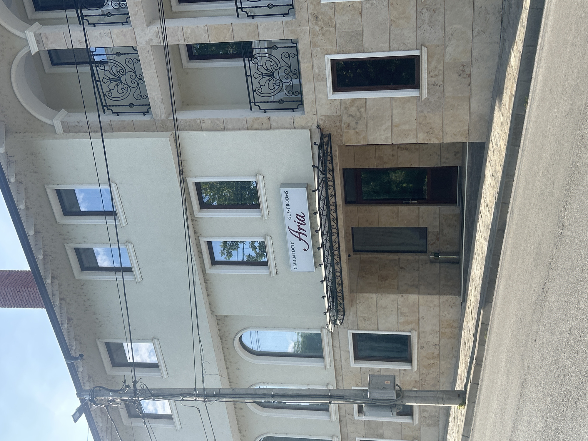 A building with a sign for "Aria Guest Rooms" stands next to a paved road. The building appears to be a multi-story structure with a beige and white facade, featuring rectangular windows and decorative black metalwork on the upper floors. The sign is mounted above the entrance and reads "СТАИ ЗА ГОСТИ Aria GUEST ROOMS." A utility pole with electrical wires is visible in the foreground. The location is likely Veliko Tarnovo, Bulgaria, based on the Cyrillic text on the sign.