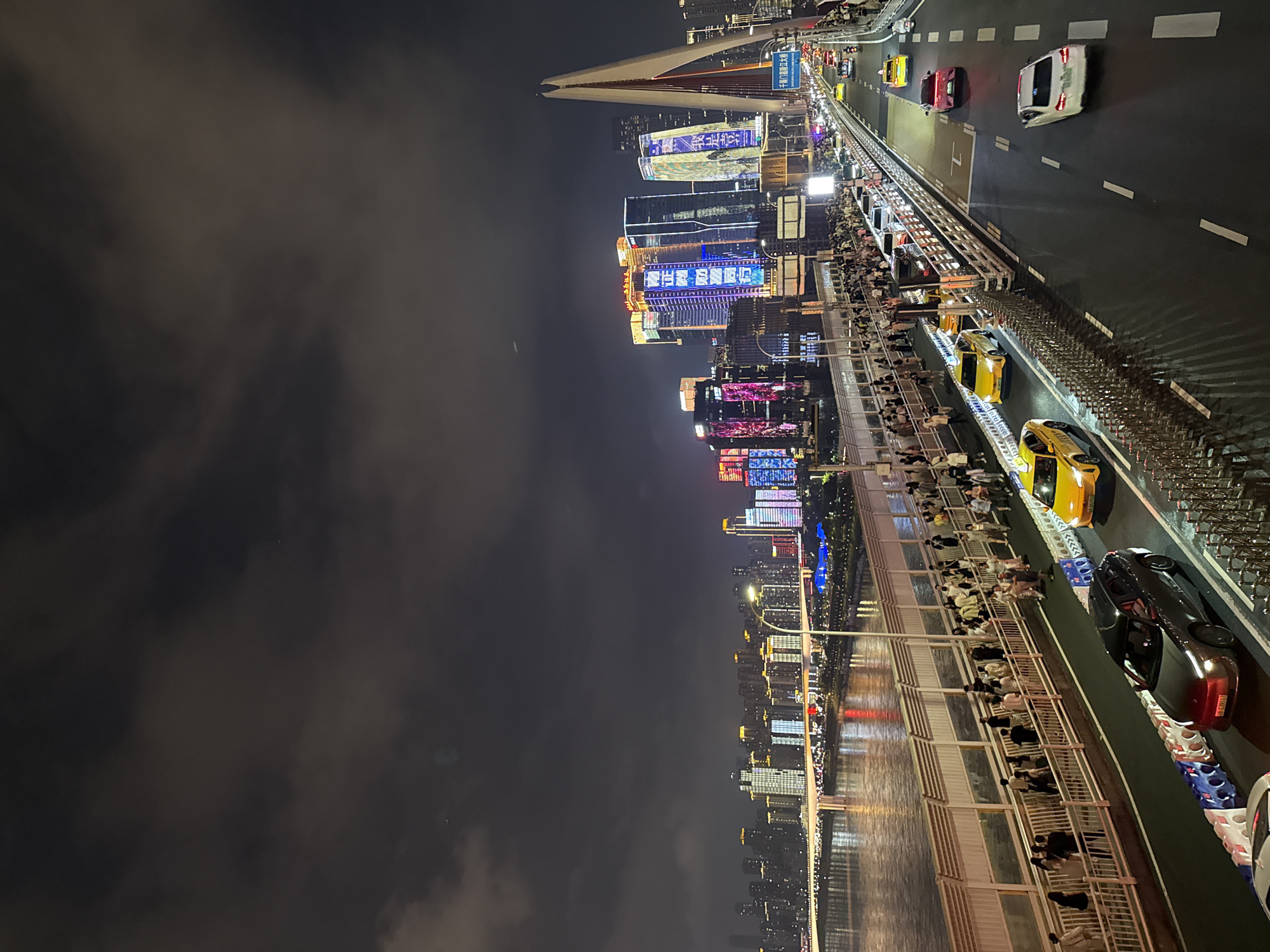 The image captures a nighttime view of the Hongyadong district in Yuzhong District, China, likely from a bridge. A wide river flows on the left, reflecting the bright lights of a cityscape that stretches across the horizon. The cityscape is dominated by numerous tall buildings, many adorned with vibrant, colorful lights, creating a dazzling urban panorama. On the right side of the image, a distinctive white cable-stayed bridge structure rises prominently, its architectural form highlighted against the dark sky. The bridge itself is a multi-lane roadway with traffic, including several yellow taxis and other vehicles, moving along it. A pedestrian walkway runs parallel to the roadway on the bridge, populated by a crowd of people who appear to be strolling and observing the scenery. The sky is dark and overcast with some diffused light.