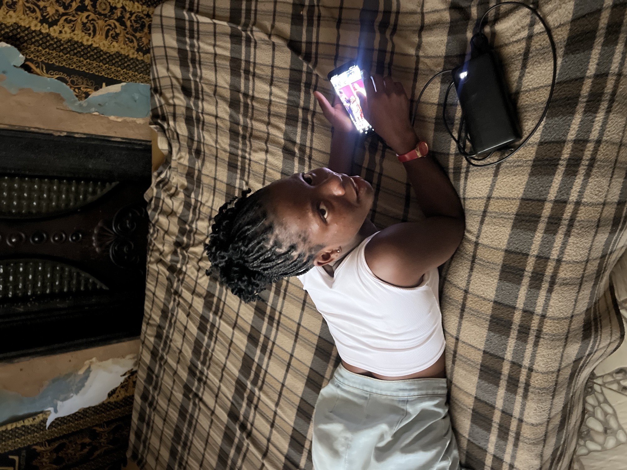 In Nkpor, Nigeria, a young woman lies on a bed with a plaid blanket, propped on her elbow and looking at her phone, which is lit up and displaying an image. She is wearing a white sleeveless top and light blue shorts. A black power bank with a charging cable is connected to her phone and lies next to her on the blanket. The background shows a wall with peeling paint and ornate wallpaper.