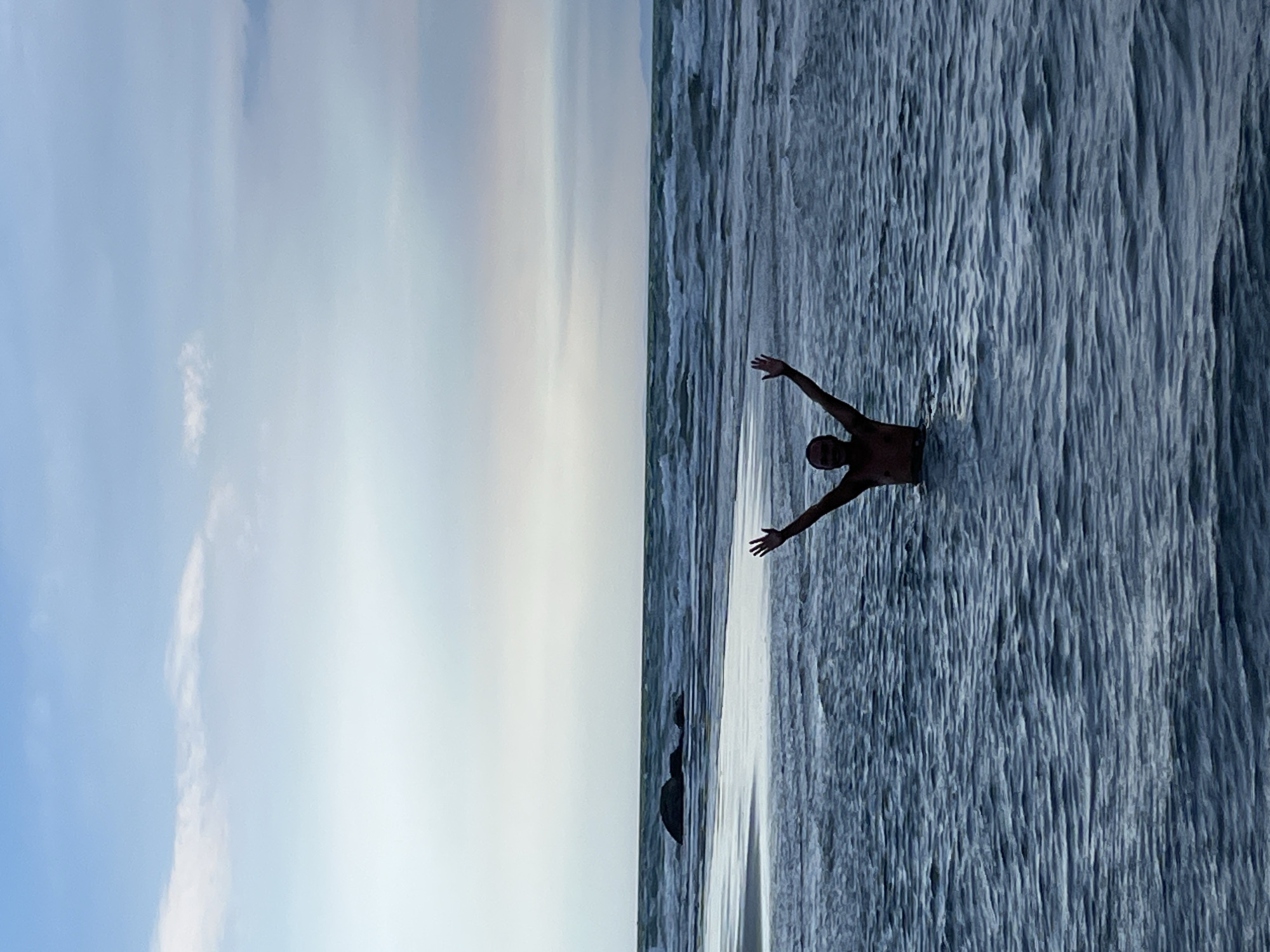 A person stands in the ocean with their arms raised in the air. The person is silhouetted against the water and sky. The water is a deep blue and appears to be choppy with small waves. In the distance, there is a beach with some rocks and the ocean continues to the horizon. The sky is a pale blue with some wispy white clouds. The scene suggests a playful or celebratory moment. The location is a beach in Mũi Né, Vietnam, a coastal resort town known for its sand dunes and beaches.