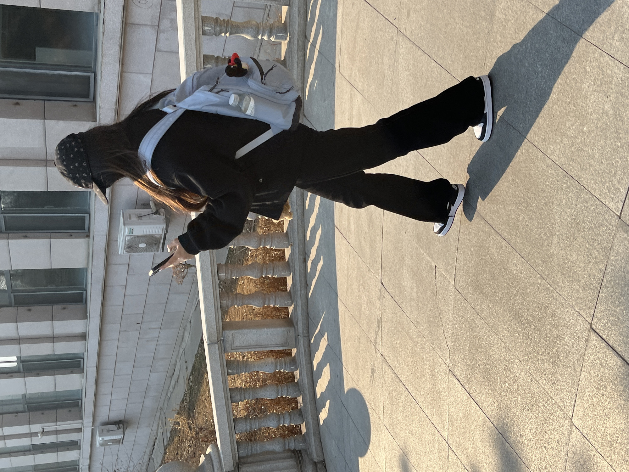 An individual is depicted walking from right to left on a tiled pedestrian path, illuminated by sunlight casting long shadows. The scene is located in Changchun City, China. The person is wearing a black baseball cap with a patterned design, a dark long-sleeved top or jacket, dark wide-leg pants, and black and white sneakers. They have long dark hair visible from beneath the cap. A light grey backpack is worn, featuring a clear water bottle in an external mesh pocket and a small black and red plush toy attached to the top. The individual is holding a dark-colored smartphone in their right hand, positioned as if viewing the screen. To their left, a light-colored stone balustrade railing with decorative pillars separates the pathway from a lower-level ground area containing sparse, dry vegetation. In the background, a large building with light-colored stone or concrete block cladding is visible, featuring multiple rectangular windows with dark frames. Two white outdoor air conditioning units are mounted on the building wall, and a small white security camera is mounted above one of the units.
