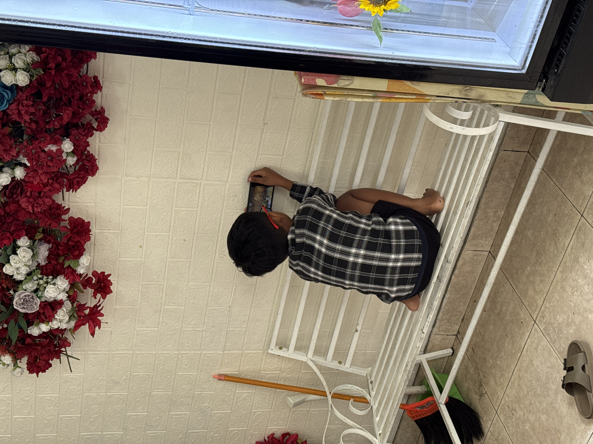 In Kuta Selatan, Indonesia, a young boy, appearing to be of Indonesian descent, sits on a white metal bench against a textured, cream-colored brick wall. He is wearing a black and white plaid shirt and dark shorts, and his feet are bare. The boy is holding a smartphone horizontally in his hands, focused on the screen as he plays a game. Red-rimmed glasses perch on his nose. To the left of the bench, a broom with a red brush and a green dustpan rests against the wall. Above the boy and bench, a large arrangement of artificial red and white flowers hangs on the wall. To the right of the boy, a patterned curtain or fabric hangs beside a glass window or door, through which a sunflower can be seen. The floor is tiled with beige squares. A single brown sandal lies on the floor in the foreground.