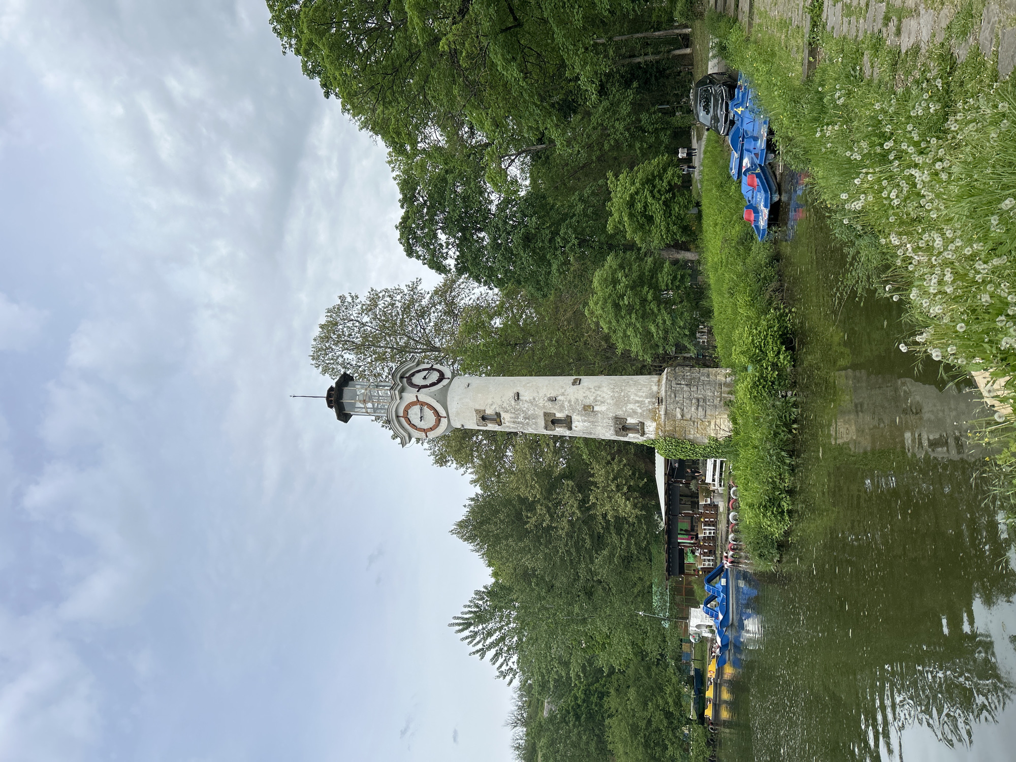The image depicts a tower, presumably a lighthouse replica, located next to a body of water, likely a canal or small lake. The tower has a weathered, white facade with a clock face visible near the top. Several life preservers are attached to the tower. Lush greenery surrounds the structure. A few paddle boats are situated along the bank near the water. The overall scene suggests a recreational area, possibly a park. The photograph appears to have been taken in Pleven, Bulgaria.