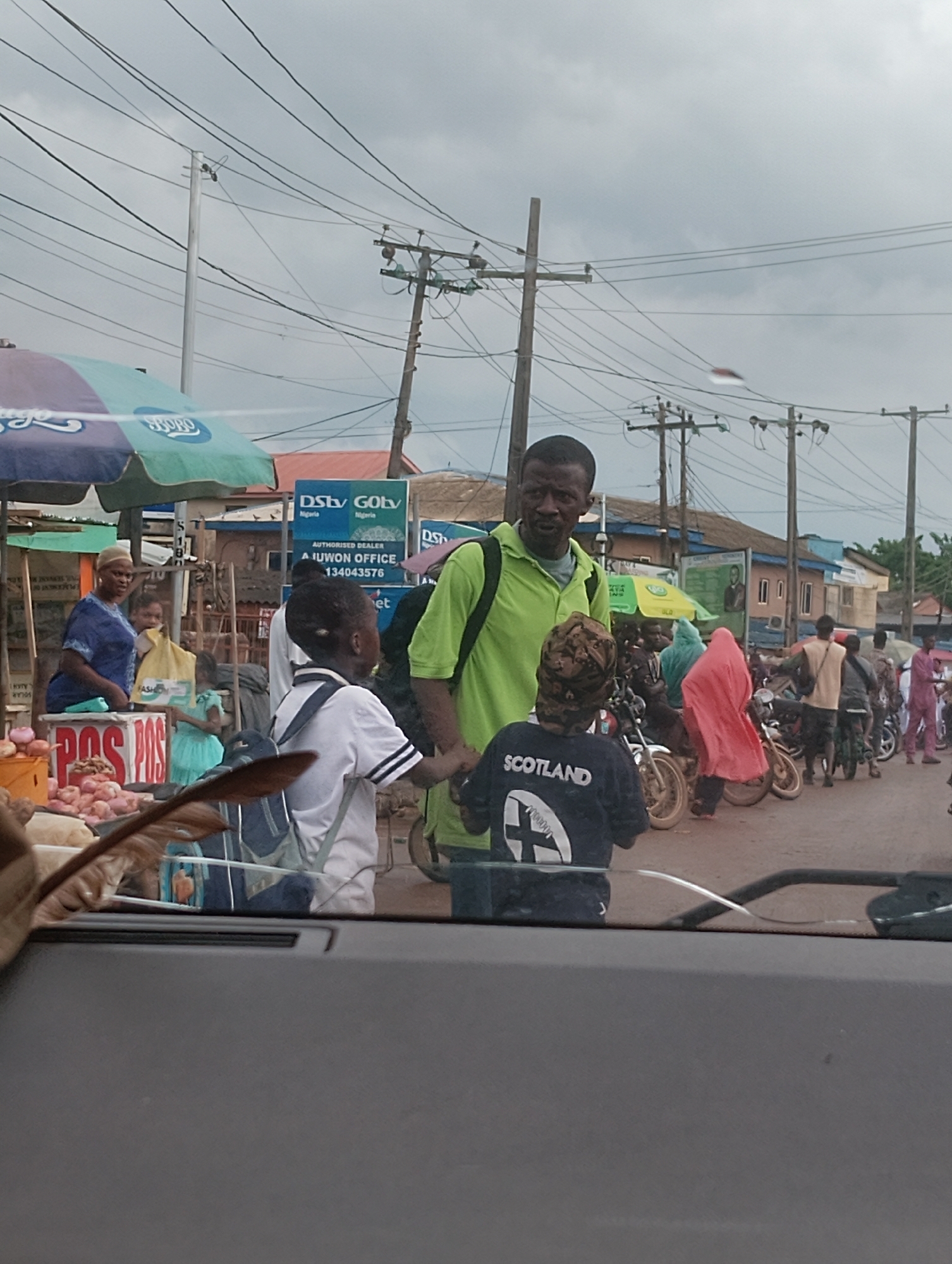This is a photograph taken on a cloudy day in Aketu-Oja, Nigeria, showing a street scene. In the foreground, a car's dashboard and windshield are visible, reflecting the scene outside. The main focus of the image is a group of people gathered on a street, likely a marketplace.
Several individuals are present. A man in a bright green shirt stands in the center, appearing to interact with a young boy in a white shirt and backpack who is facing away from the camera. To the left, a woman in a blue and white patterned outfit stands behind a stall laden with onions and other produce. Another young person in a turquoise dress is also visible near the stall.
Behind these individuals, several motorcycles are parked, indicating a mode of transportation common in the area. People are seen standing around the motorcycles. Signs for "DStv" and "GOtv" are visible on a building in the background, suggesting a commercial area. Electrical poles and wires crisscross the sky, typical of urban infrastructure. The overall impression is a bustling street scene with elements of commerce and daily life.