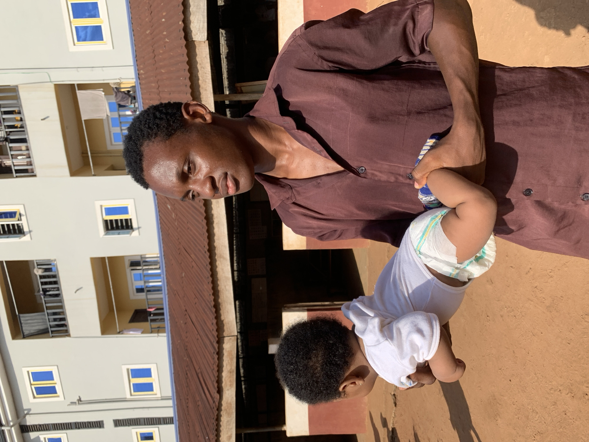A medium outdoor shot depicts an African man holding a baby in what appears to be Ukwu-Abwa, Nigeria. The man is wearing a dark brown, button-down shirt. The baby is wearing a white shirt and diaper. The man and baby are in front of a light gray building. A red corrugated metal roof is in the background.