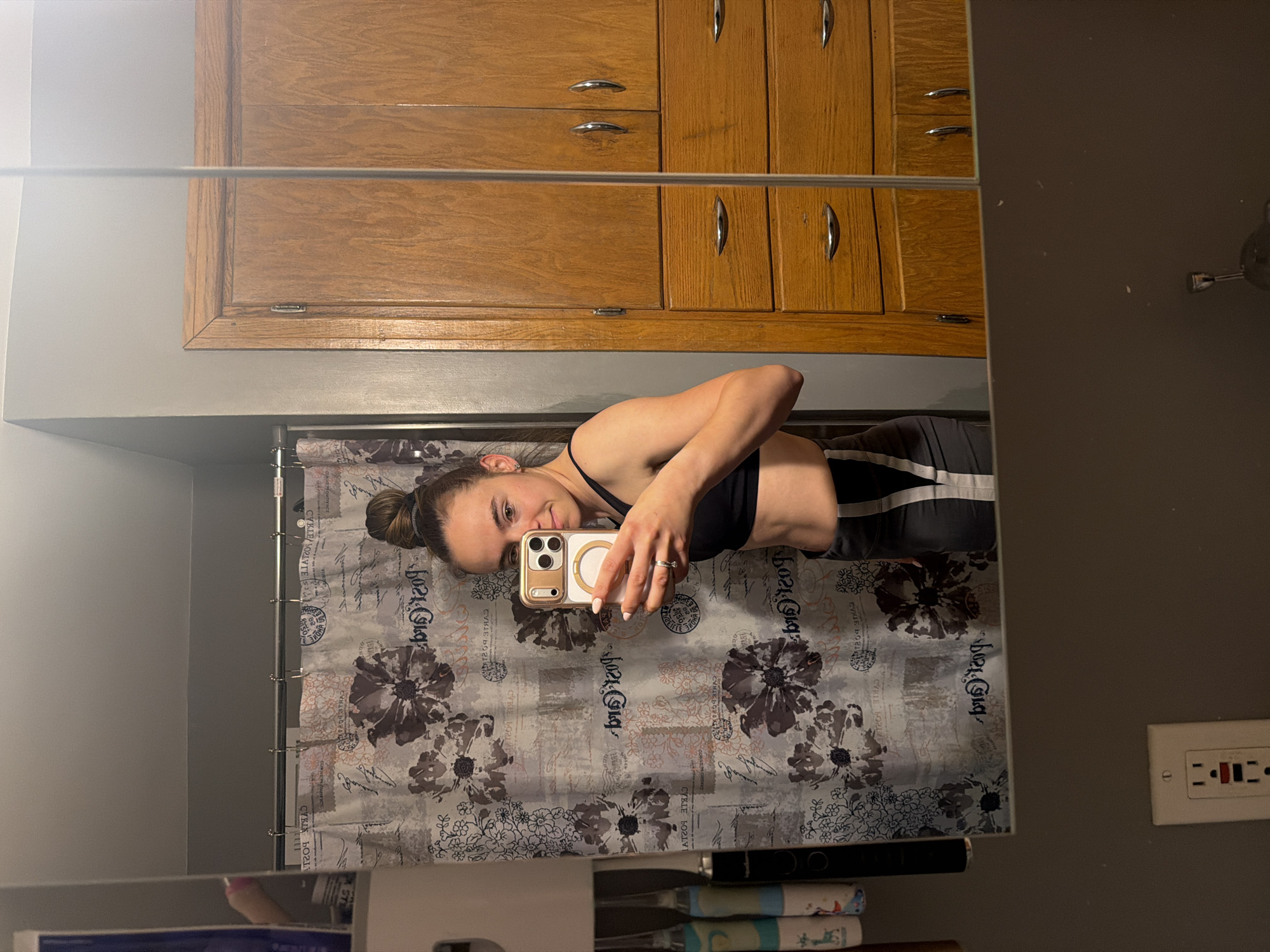 The image shows a person reflected in a mirror. The person is holding a phone, likely taking the photo. They have a bun hairstyle and are wearing workout clothes. The background includes a bathroom with a patterned shower curtain and wooden cabinets. An electrical outlet is visible on the right wall of the bathroom. There is no identifiable location in the city of Reedsburg, United States of America.