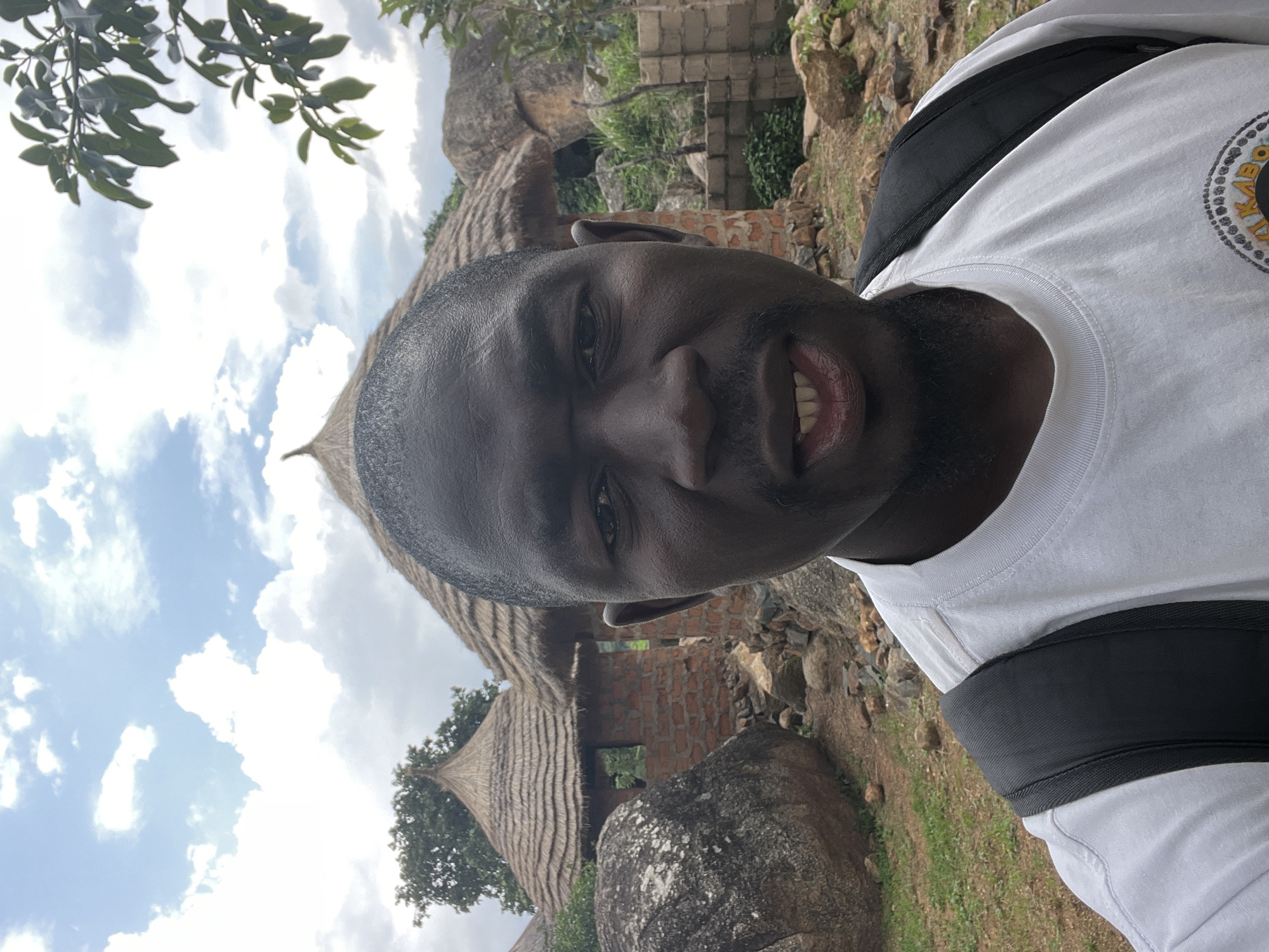 The image is a selfie taken outdoors in front of traditional thatched-roof huts in the city of Jos, Nigeria. A Black man wearing a white t-shirt and a black backpack is the central subject, positioned in the foreground. His face is partially obscured by the close-up framing of the selfie. Behind him, the huts, constructed with red brick and topped with straw, are visible, along with large rocks and scattered vegetation. The sky above is cloudy with patches of blue. The overall environment appears to be rural or semi-rural.