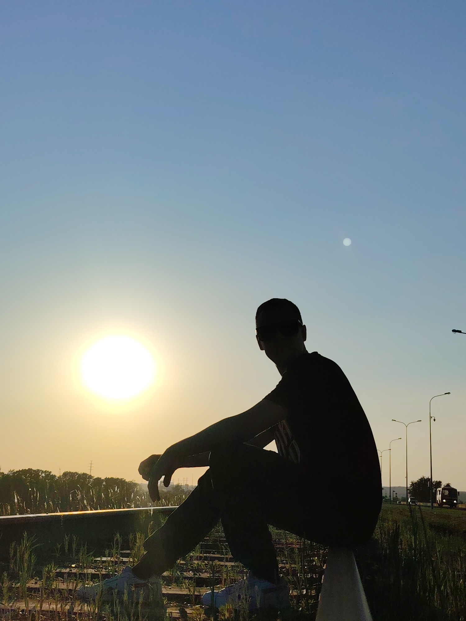 The image depicts a silhouetted figure sitting on a metal rail against a backdrop of a bright sunset or sunrise. The location appears to be an open area possibly in or near a city. A line of streetlights stretches into the distance on the right side of the image, with traffic visible on a road alongside. There are trees and fields to the left, and the presence of vegetation around the rail suggests a rural or suburban setting. Given the prompt, this scene is likely located in or near the city of Chaikovsky, Russia.