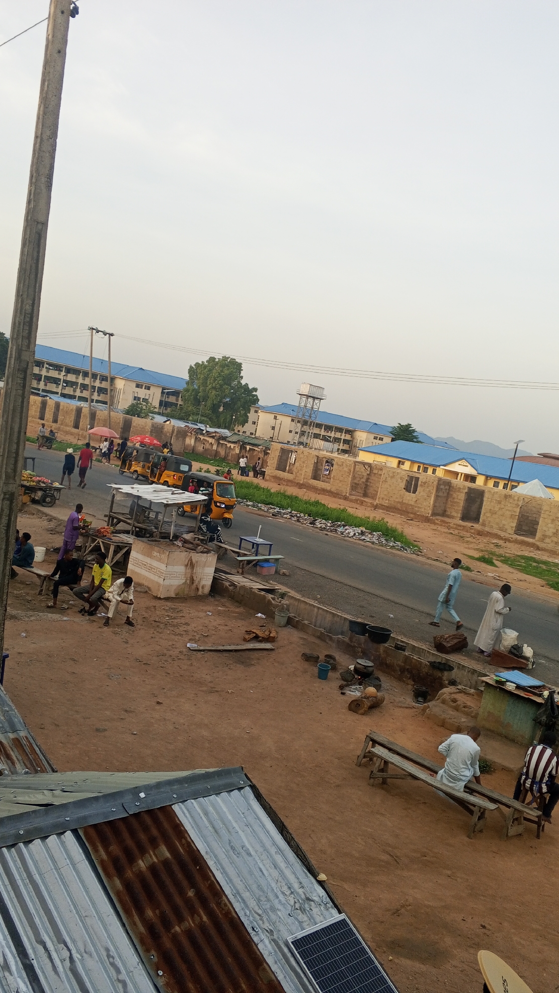 An outdoor, eye-level, wide shot captures a street scene in a city, likely in Africa, given the presence of auto-rickshaws. The time of day appears to be late afternoon or early evening, with soft, overcast lighting. The environment is predominantly earth-toned, with unpaved ground and partially constructed buildings made of tan bricks. In the foreground, a corrugated metal roof is visible, with a solar panel on the right side.
Several individuals are present. On the left, a cluster of people are gathered around market stalls and sitting on wooden benches. One person in a purple shirt stands near a stall. Further down the street, three yellow auto-rickshaws are driving along a paved road. Several more people are walking and standing on the side of the road. On the right side of the road, two men are walking. One man in a pale blue outfit is moving towards the left, and another man in a white robe is moving towards the right. Two other men are sitting on a wooden bench on the right side of the street, one in a white shirt and the other in a striped shirt.
In the background, a multi-story building with blue roofs and tan walls is visible, along with other structures that appear to be residential or institutional. A water tower stands behind some of these buildings. The street is lined with a low wall, and vegetation is sparse, with some trees and grass visible in the distance. The overall impression is of a bustling, everyday street in a developing urban area.