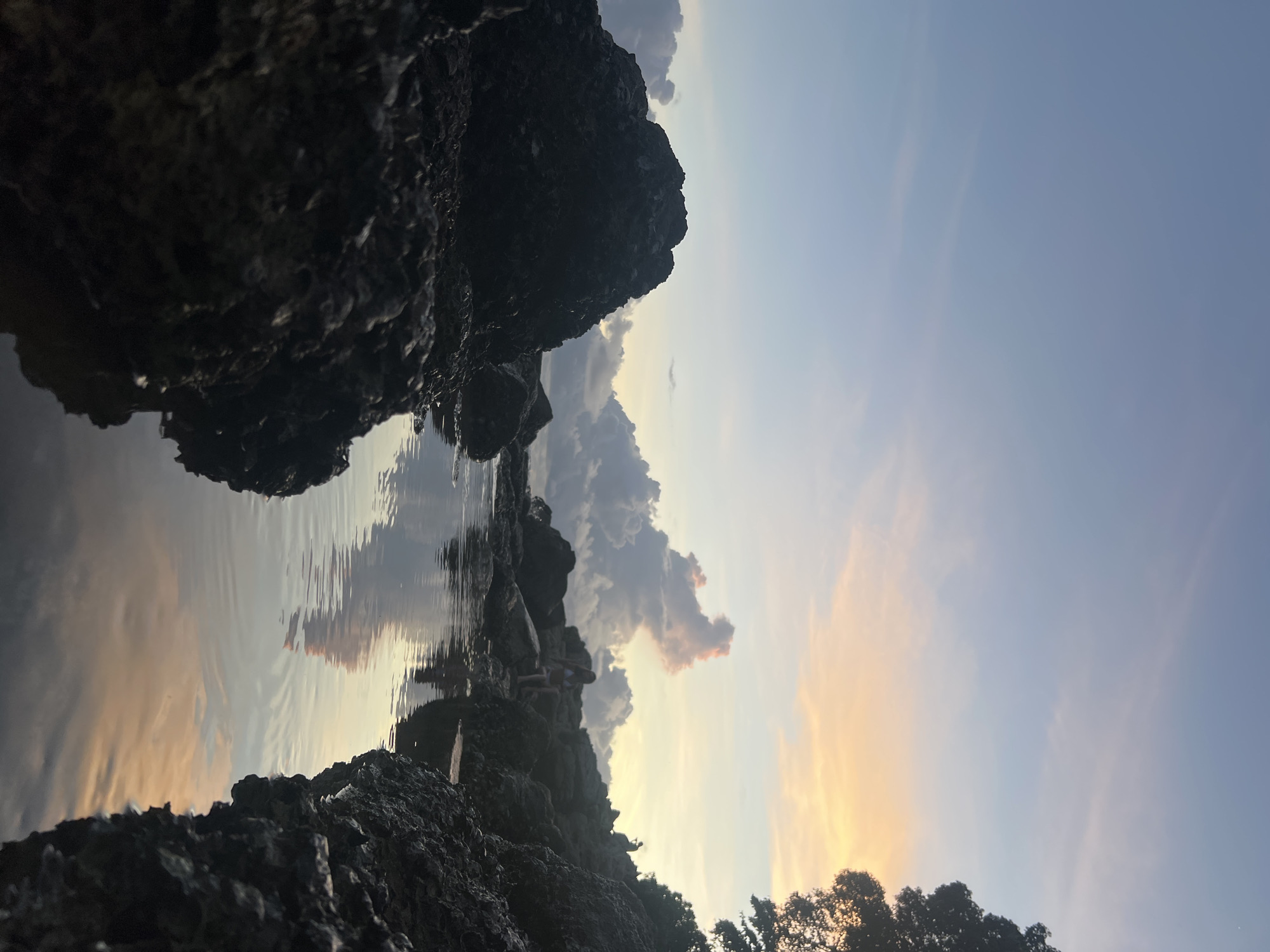The image depicts a rocky coastal scene at dusk, likely in the vicinity of Choeng Thale, Thailand. In the foreground, dark, jagged rocks frame a calm pool of water. The water reflects the soft hues of the sunset sky, which is a gradient of pale blue, pink, and orange. In the middle ground, several people are visible on the rocky shoreline. One individual, wearing a light-colored bikini, is seated on a rock. Further back, another person stands, silhouetted against the horizon. A dense tree is present on the right side of the image, its dark branches silhouetted against the colorful sky. The overall atmosphere is serene, capturing the tranquil beauty of a tropical sunset.