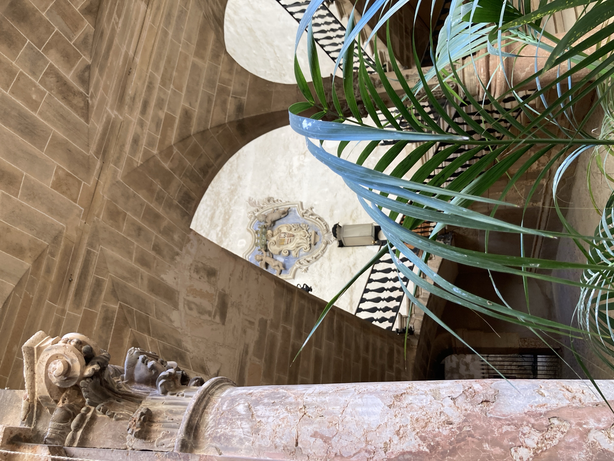 A low-angle, medium shot captures an interior courtyard in Palma de Mallorca, Spain. In the foreground, a large, decorative stone column with intricate carvings is visible on the left. Behind it, the underside of a stone arched ceiling is illuminated. In the middle ground, a staircase with ornate black balusters curves upwards. A crest with a coat of arms is mounted on a light-colored wall in the background, positioned between two arches. Green palm leaves partially obscure the view of the staircase and the wall. The scene is characterized by the interplay of light and shadow on the stone architecture and foliage.