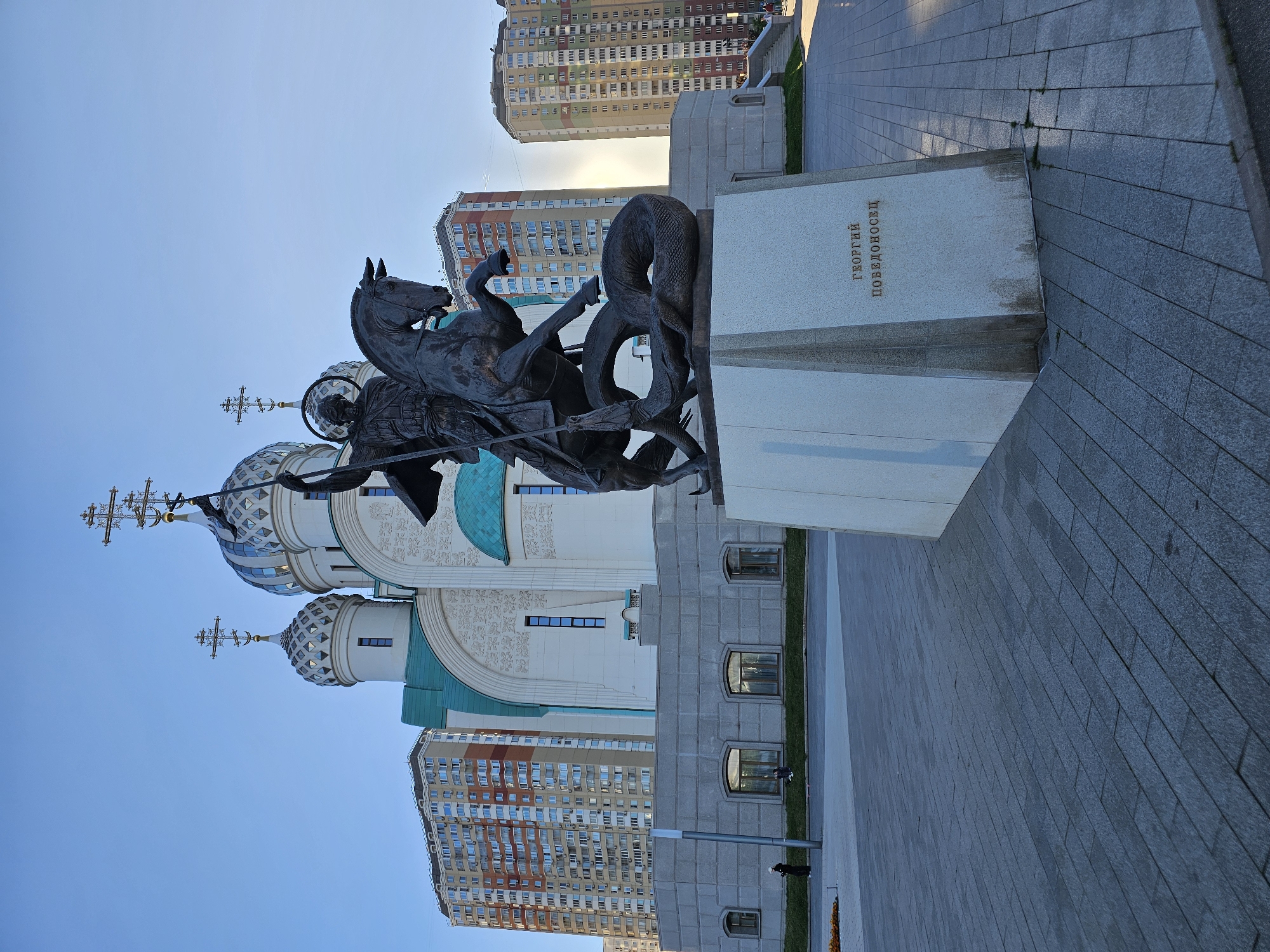 A statue of Saint George slaying a dragon is prominently featured in the foreground, with the horse and rider dynamically posed. Behind the statue, a church with distinctive blue and white domes and crosses is visible, its architecture suggesting a place of worship. To the right of the statue, a light-colored pedestal bears Cyrillic text that reads "ГЕОРГИЙ ПОБЕДОНОСЕЦ" (George the Victorious). In the background, modern, multi-story residential buildings with colorful facades dominate the skyline. The scene is set in an urban environment, likely a public square or plaza, with paved walkways and grassy areas. The clear blue sky suggests it is daytime, possibly late afternoon, with the sun casting long shadows. The overall atmosphere is one of civic monumentality and religious presence, set against a backdrop of contemporary urban development.