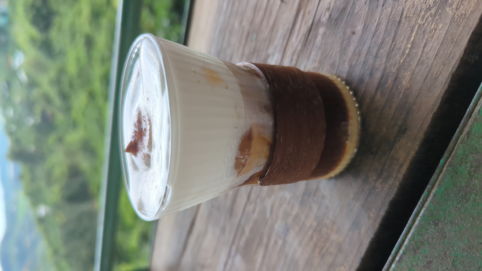 A close-up, eye-level shot focuses on a layered beverage in a clear glass, resting on a weathered wooden table. The drink features distinct layers: a light tan bottom, a dark brown middle, and a creamy white foamy top. A small pile of reddish-brown powder, likely cinnamon or cocoa, is dusted onto the center of the foam. The glass is encircled by a textured dark brown band. The background is softly blurred, revealing lush green, hilly terrain under a hazy sky, suggesting a mountainous or elevated rural setting, potentially within Thôn 5, Vietnam, known for its natural landscapes. A green metal railing runs horizontally in the mid-ground, separating the table from the vista. The overall lighting is natural and diffused, creating a serene ambiance.