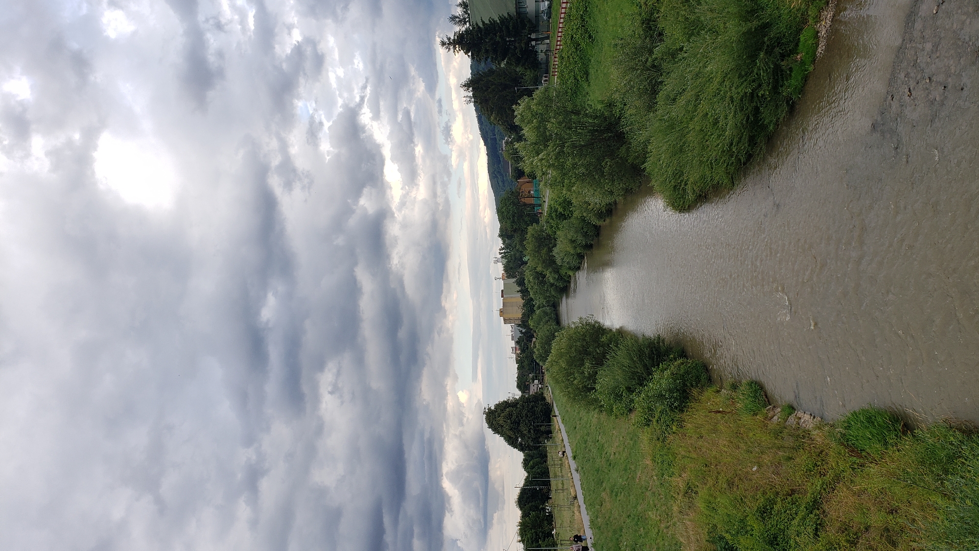 An overcast sky with patches of bright sunlight looms over a scenic view of the Sekčov River in Prešov, Slovakia. The river flows through a lush green landscape, bordered by grassy banks and dense foliage on either side. In the background, the city skyline features a cluster of buildings, including a prominent yellow structure. A paved pathway runs along the left bank of the river, where several individuals are visible walking, enjoying the serene environment. The overall impression is one of a tranquil day in a picturesque urban setting, with nature and city life coexisting harmoniously.