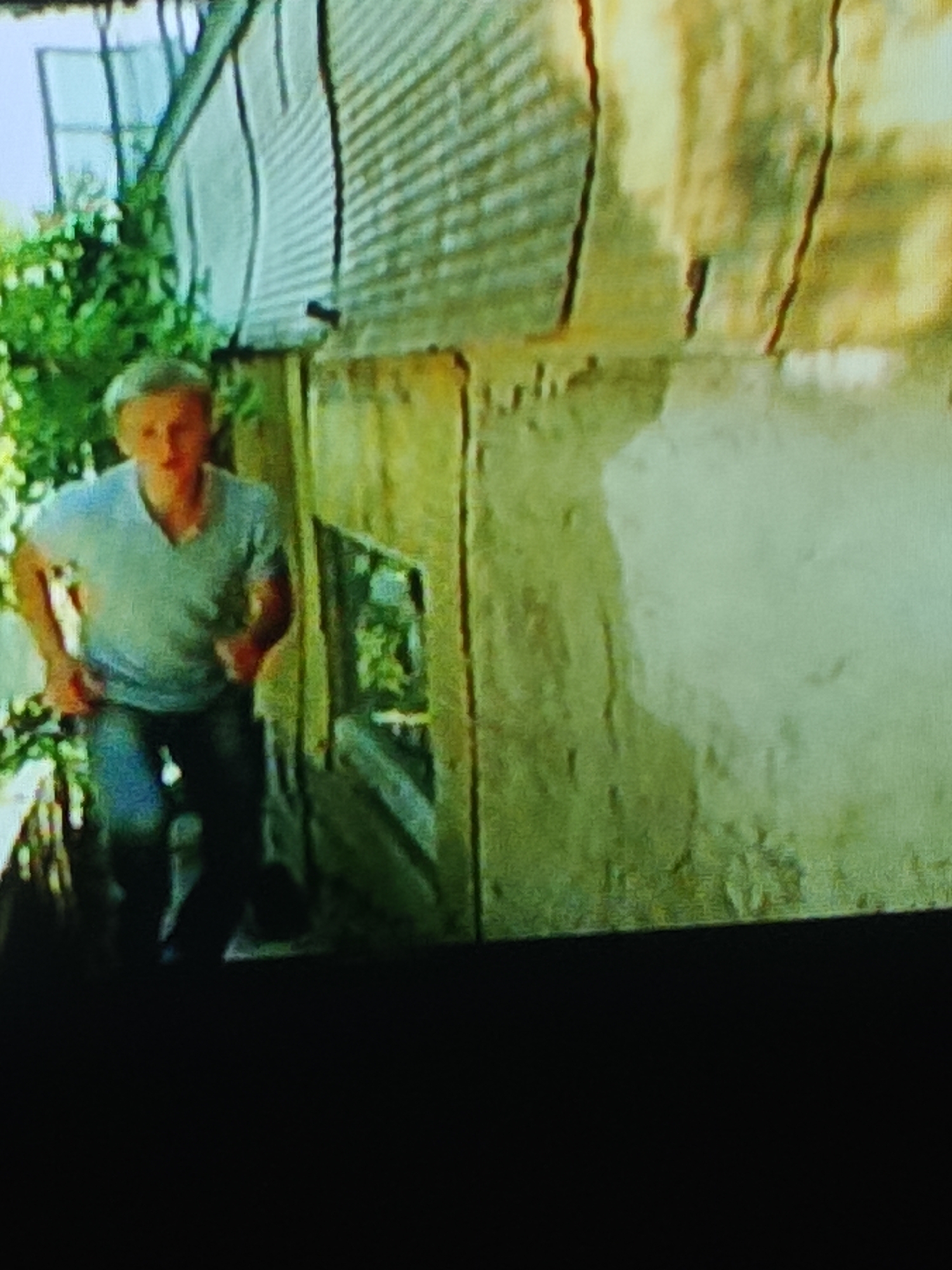 A young man with blonde hair, wearing a light blue v-neck t-shirt and dark pants, is running towards the left side of the frame. He appears to be in an outdoor setting, with a weathered, corrugated metal structure to his right and lush green foliage to his left. The lighting suggests it is daytime. The man's body is angled, suggesting movement. The background consists of the textured metal wall of a building and some greenery. There is no visible indication of the city of Glazov, Russia, within the image itself.