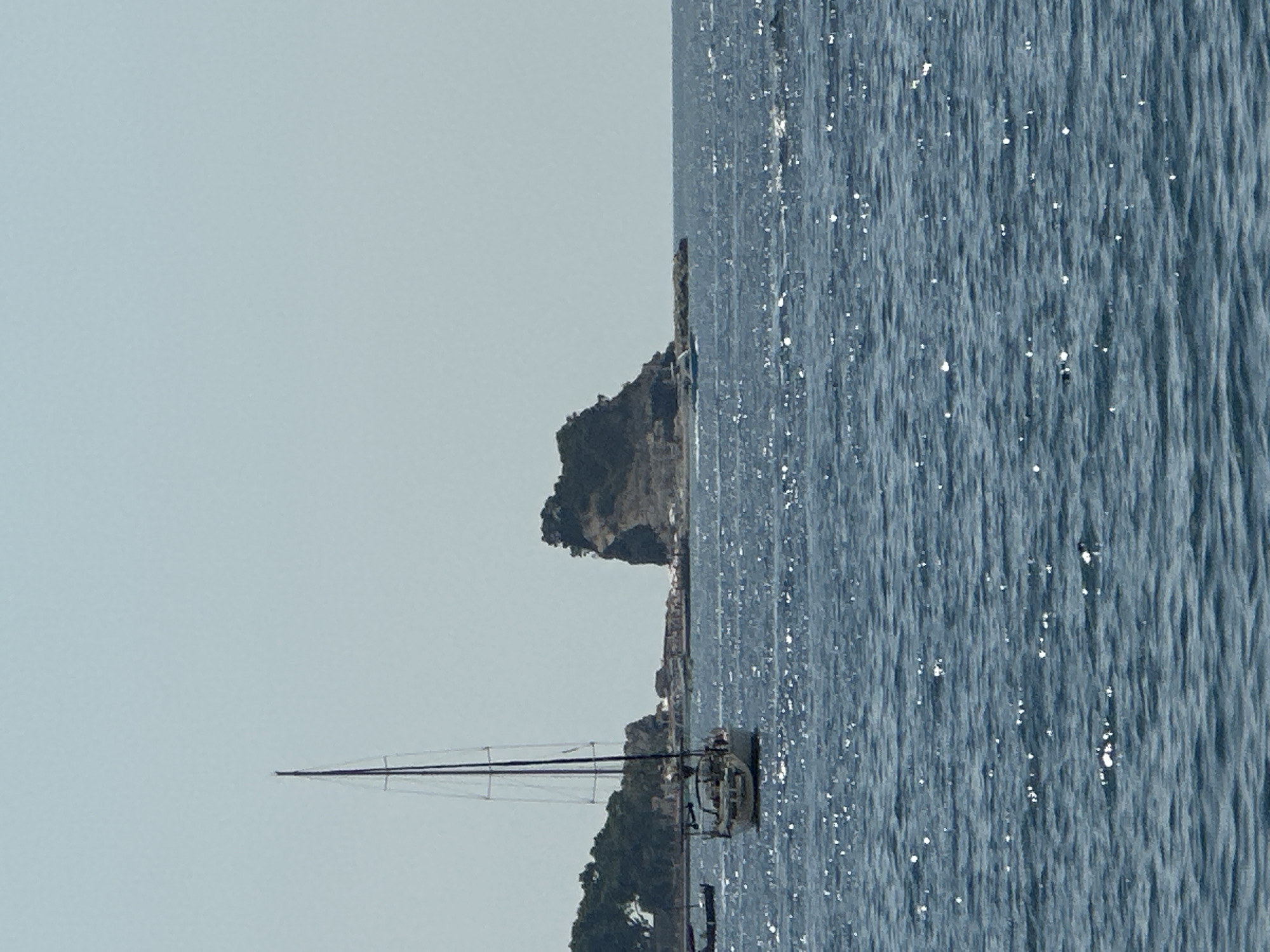 The image shows a view of the Adriatic Sea from Budva, Montenegro. In the foreground, the sea is blue and choppy with sunlight glinting off the surface. A white sailboat with a tall mast is visible in the middle ground, to the left of the frame. In the background, a rocky island, St. Nicholas Island, is a prominent feature. Trees cover the slopes of the island, and a few buildings are visible on the coastline near the island. The sky is a pale blue and clear. The overall scene is one of a calm, sunny day at sea.