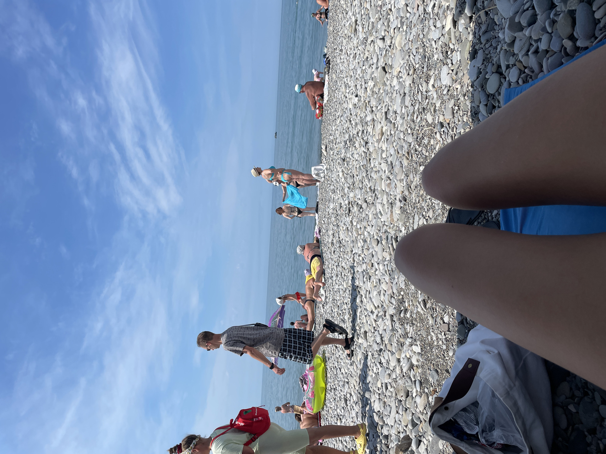 A first-person perspective shot of a beach shows a pebble shore leading to the ocean under a partially cloudy blue sky. In the foreground, a person's legs are visible, lying on a blue towel with a white bag next to them. Several people are scattered across the beach, sunbathing and walking. Some individuals are in swimwear, while others wear casual clothing. The ocean appears calm, with a few distant boats on the horizon. The overall scene depicts a relaxed day at the beach.
