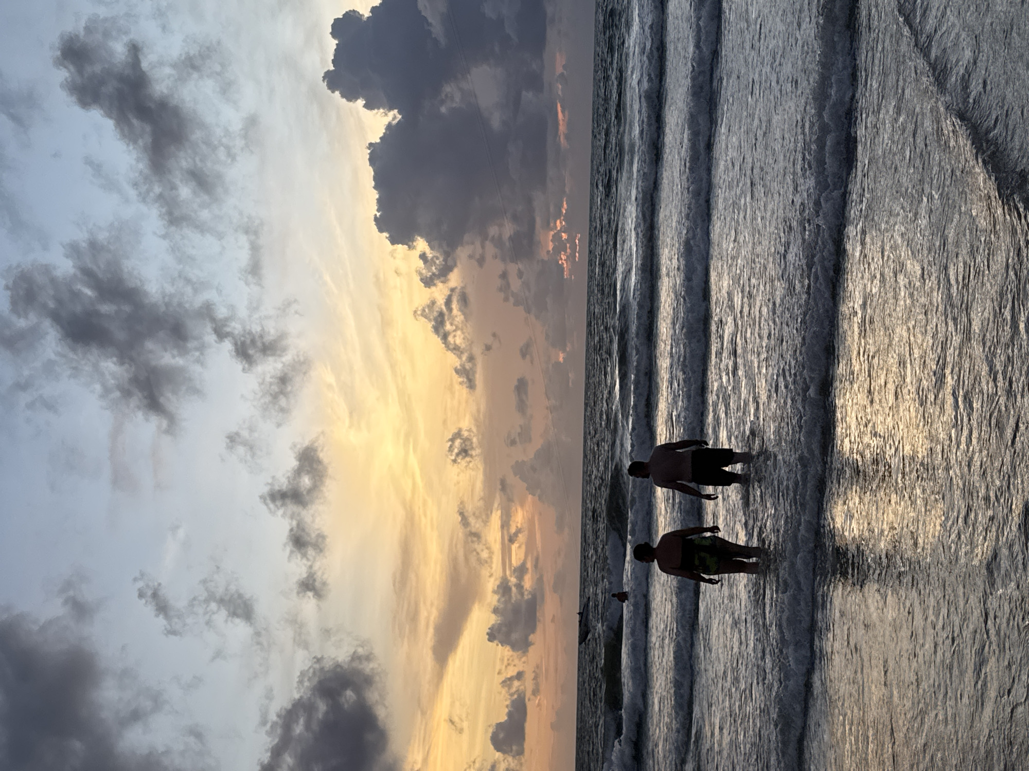 Two individuals, silhouetted against the setting sun, wade into the ocean at Patong Beach, Thailand. The sky is a canvas of dramatic clouds, streaked with hues of orange, yellow, and gray, indicative of a sunset. Waves gently roll onto the shore, their crests catching the fading light. The water is calm, with ripples reflecting the twilight sky. One person wears a patterned swimsuit, while the other is in dark shorts. A small boat is visible in the distance, further out at sea. The overall scene depicts a tranquil evening on the beach.